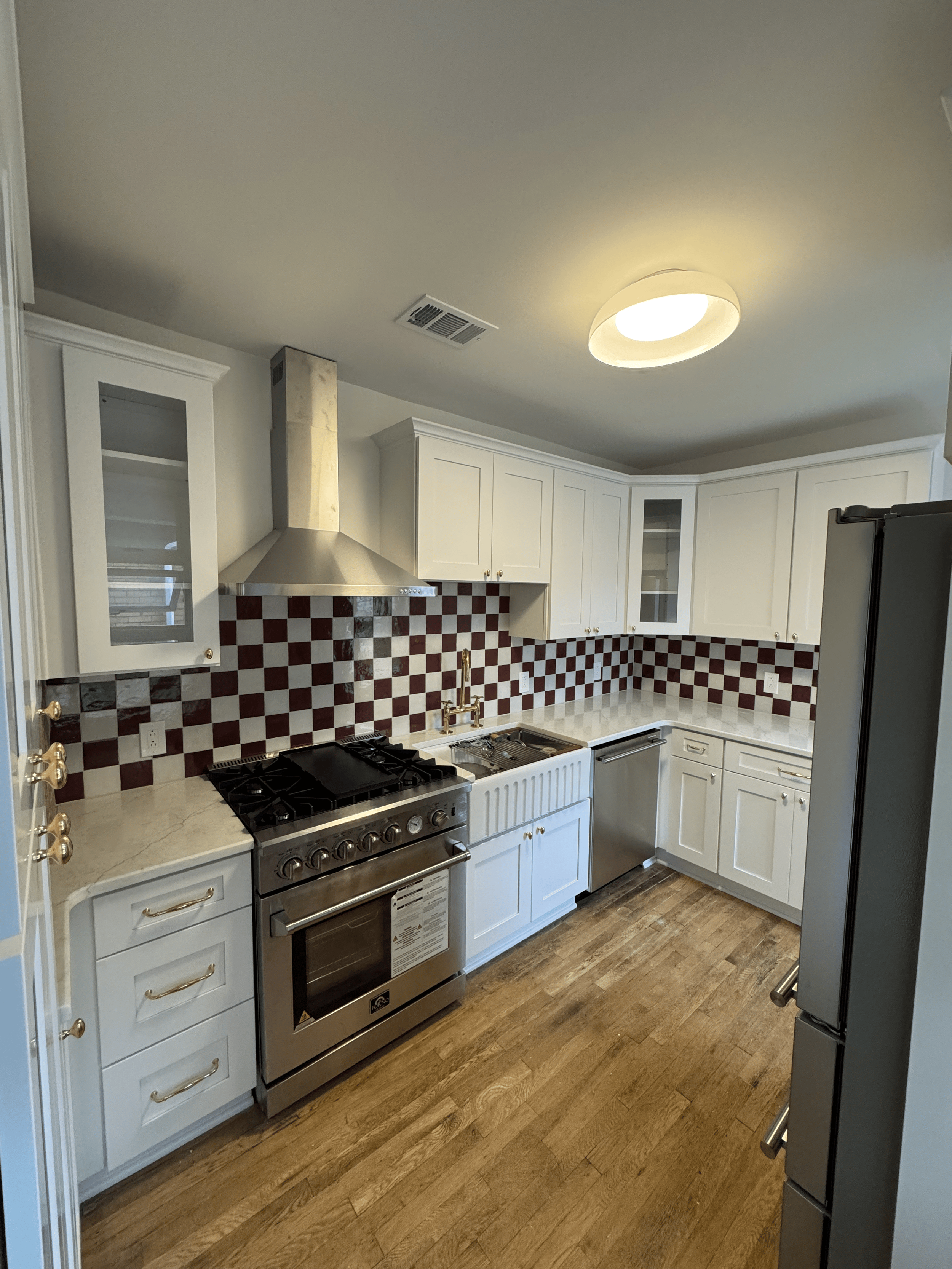A modern kitchen with white cabinets, a stainless steel oven, a stove, a range hood, a black refrigerator, and a wood floor, with a ceiling light fixture.