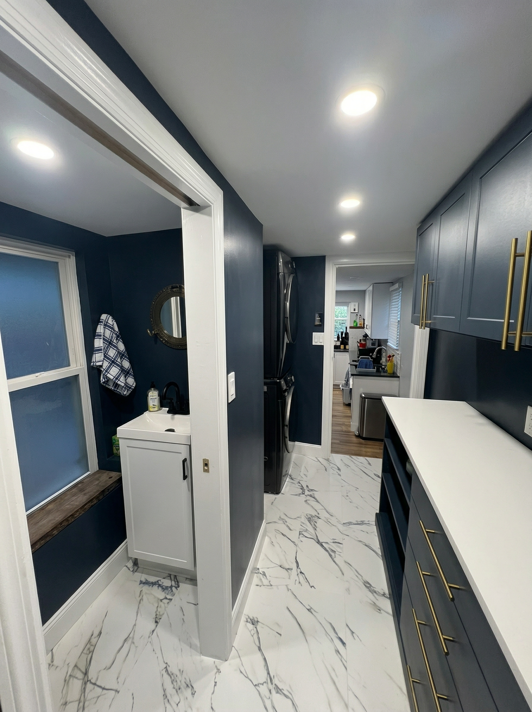 Modern kitchen with navy blue cabinets with gold handles, white marble floor with gray veining, white countertops, wall-mounted washer and dryer, and a small white sink with a mirror and towel hanging nearby.