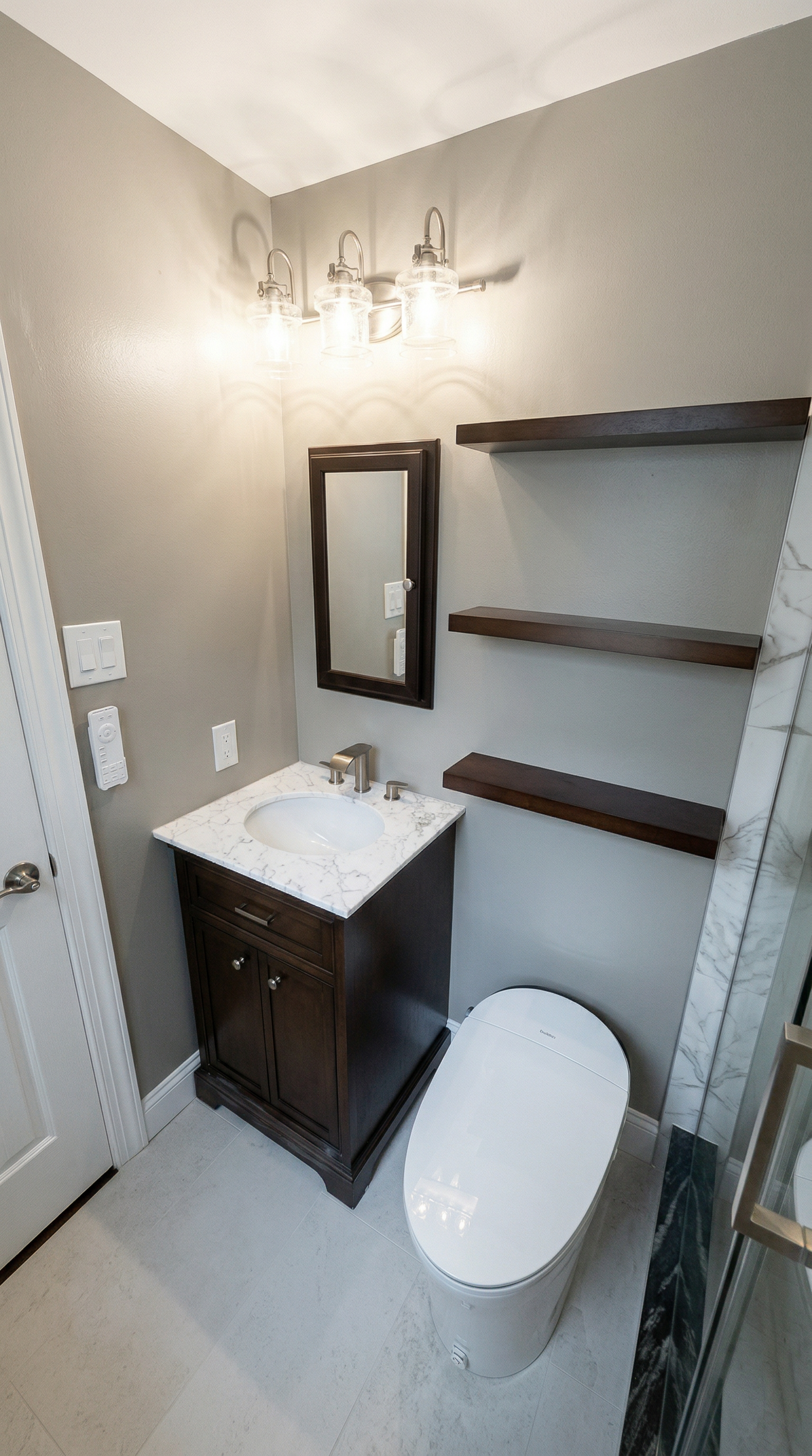 A small bathroom with a dark wood vanity and marble countertop, a mirror, bookshelf, wall-mounted light fixture, and a modern toilet.