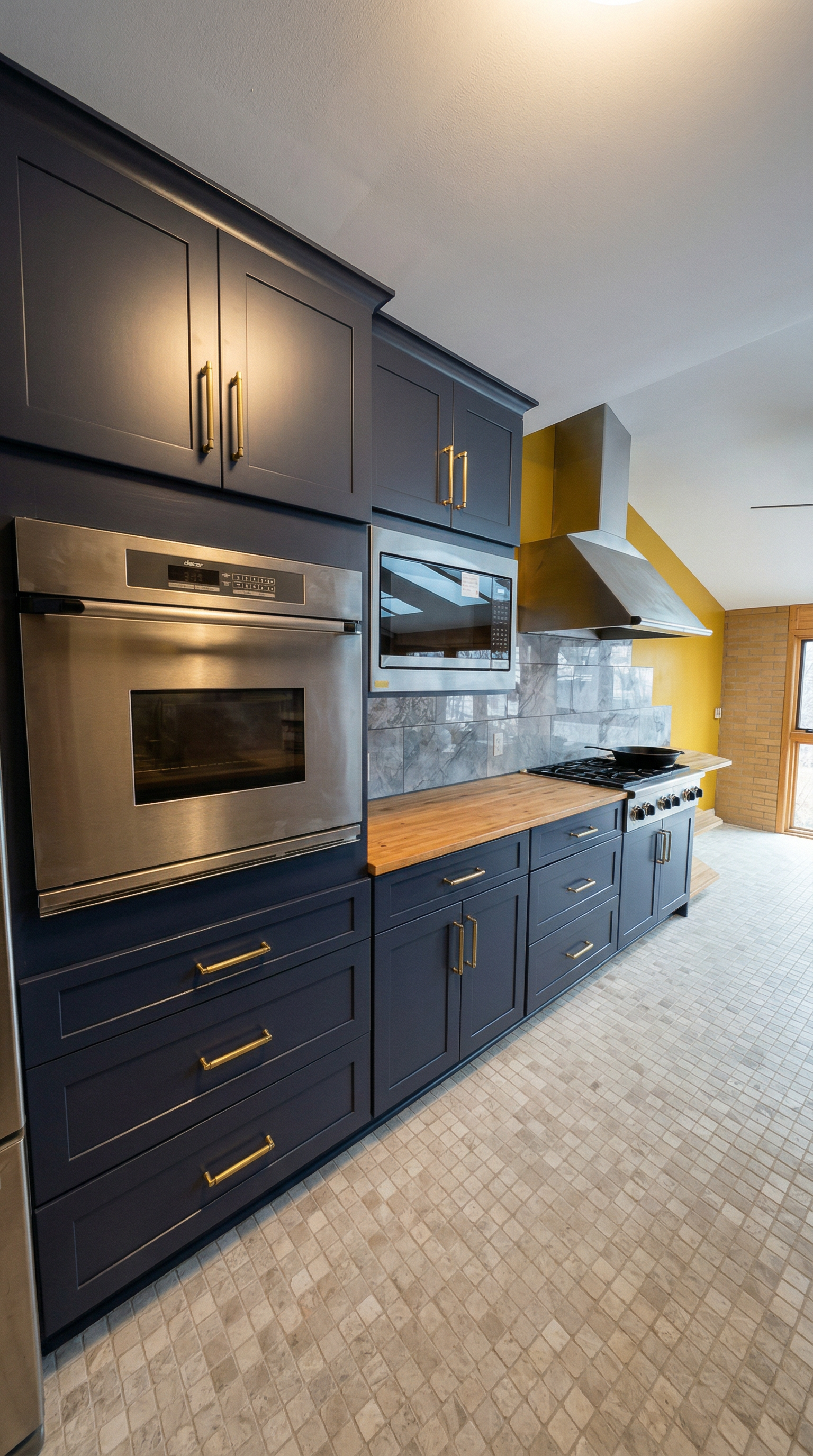 A modern kitchen with dark blue cabinets, stainless steel oven and microwave, a wooden countertop, gray marble backsplash, and yellow walls. There is a stove with a black frying pan and a vent hood above it. The floor is tiled with small square mosaic tiles.