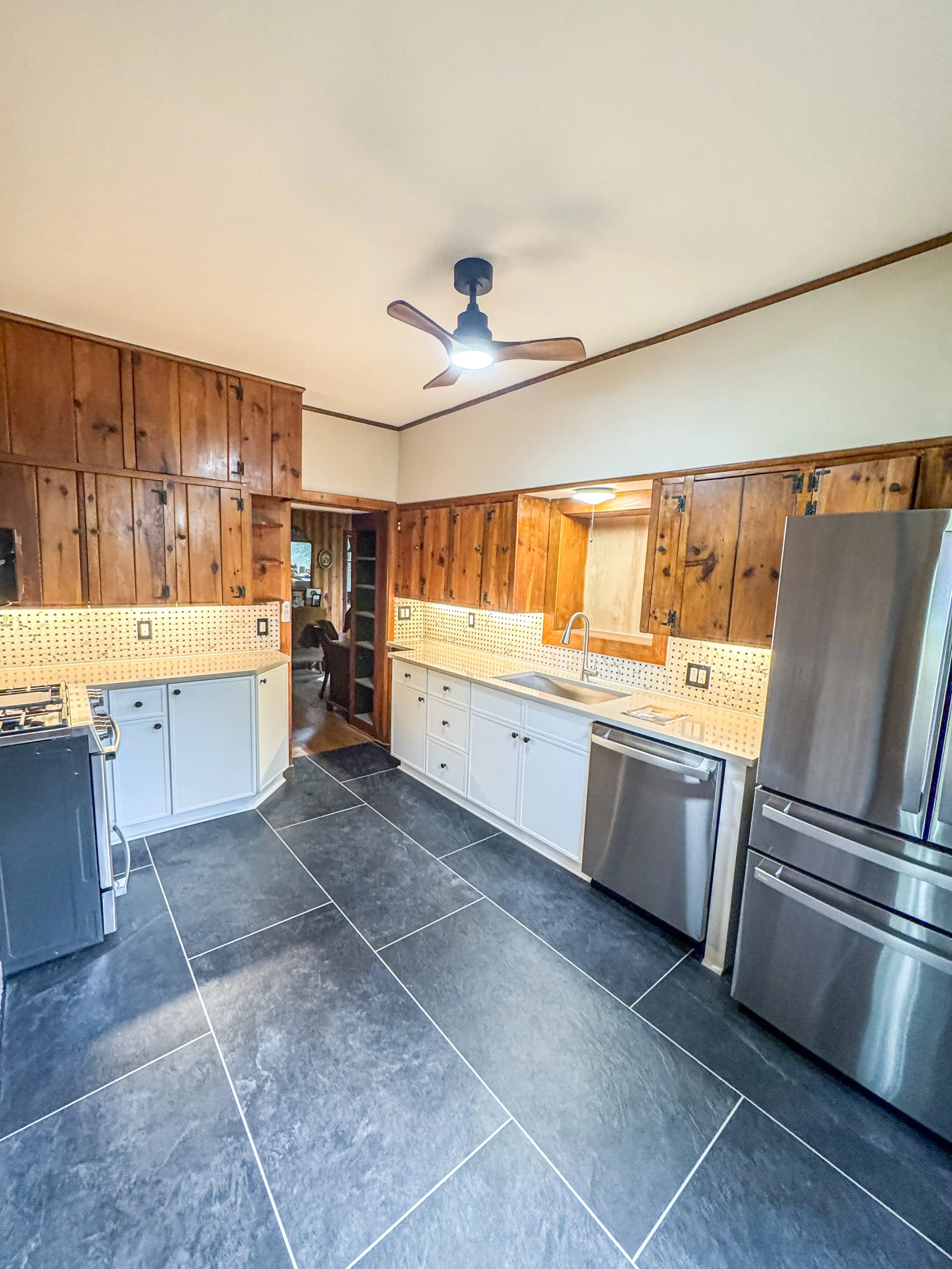 Kitchen with wooden upper cabinets, white lower cabinets, black tiled floor, stainless steel refrigerator, and ceiling fan with light.