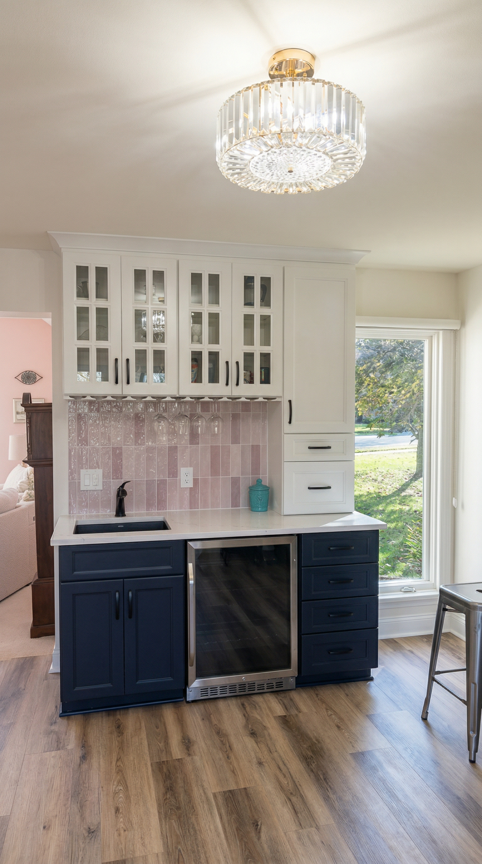 Modern kitchen with a blue lower cabinet, white upper cabinets with glass doors, pink tiled backsplash, small sink, and a wine cooler. Large window with a view of a green yard, and a crystal ceiling light.