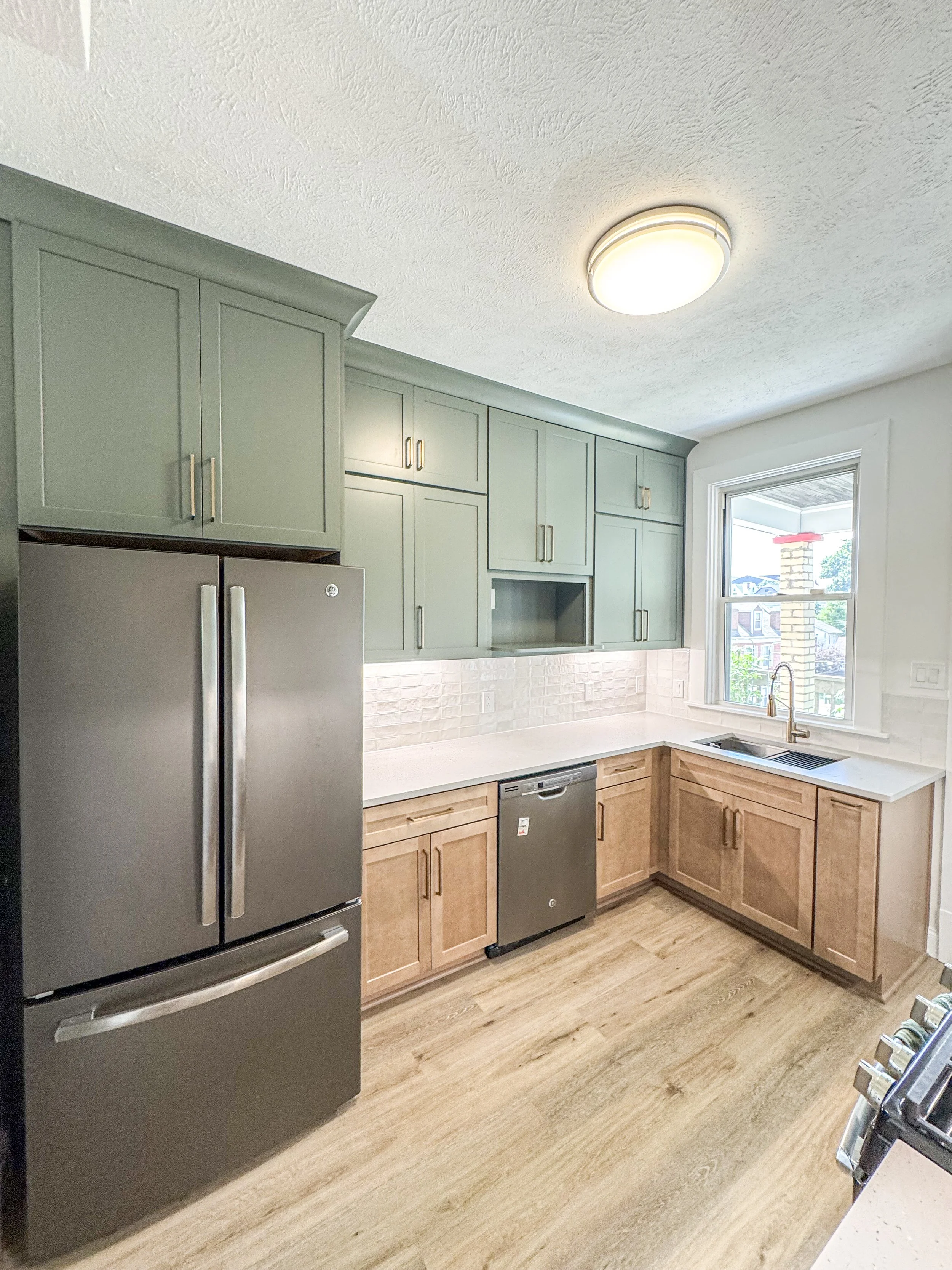 Modern kitchen with green upper cabinets, light wood lower cabinets, stainless steel refrigerator and dishwasher, large window with view of neighborhood, wood flooring, and white textured ceiling with flush-mount light.