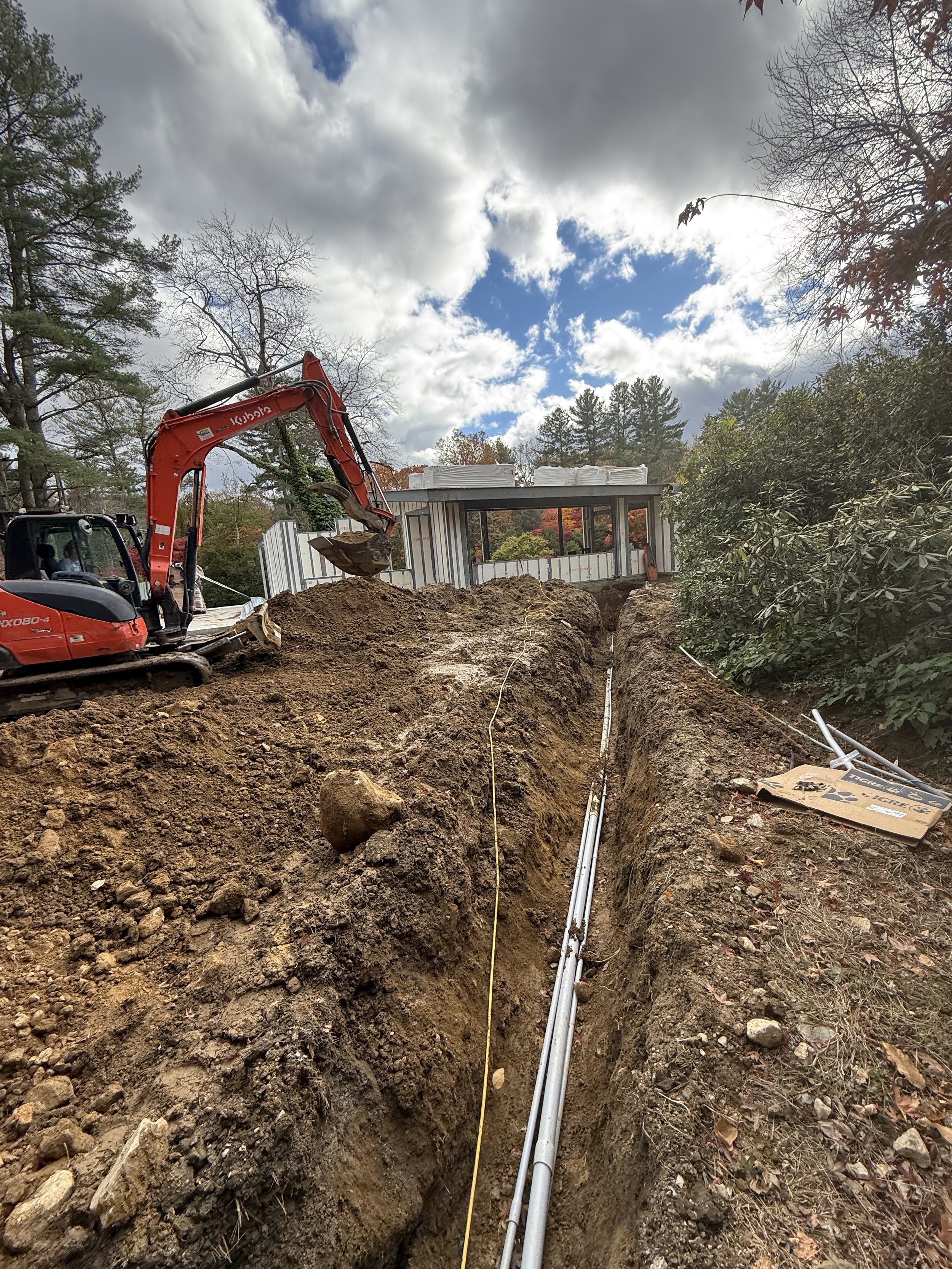 Construction site with a partially built structure, an excavator, a long pipe running in a trench, trees, and a cloudy sky.