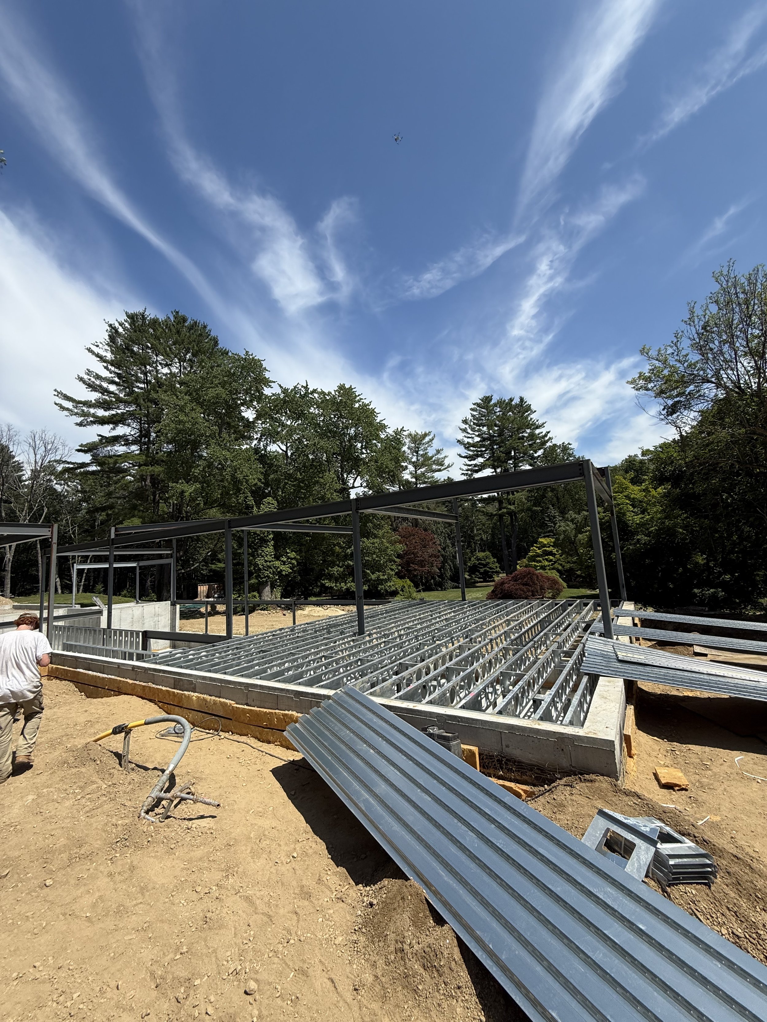 Construction site with metal framework for a building, surrounded by trees and a partly cloudy sky. Worker visible on the left.