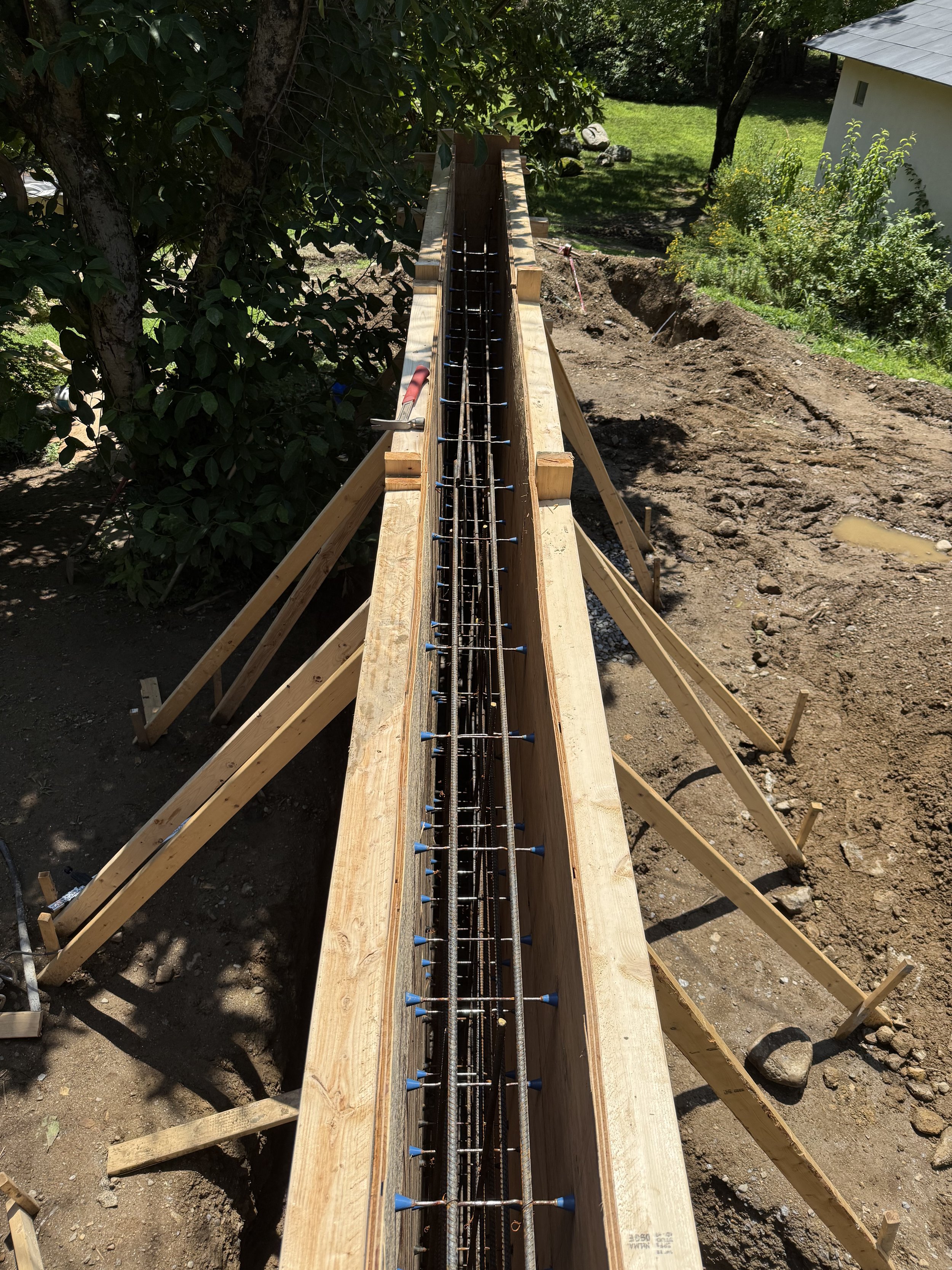 Construction site showing a wooden form for concrete with rebar inside, set on a dirt ground with trees, grass, and a house in the background.