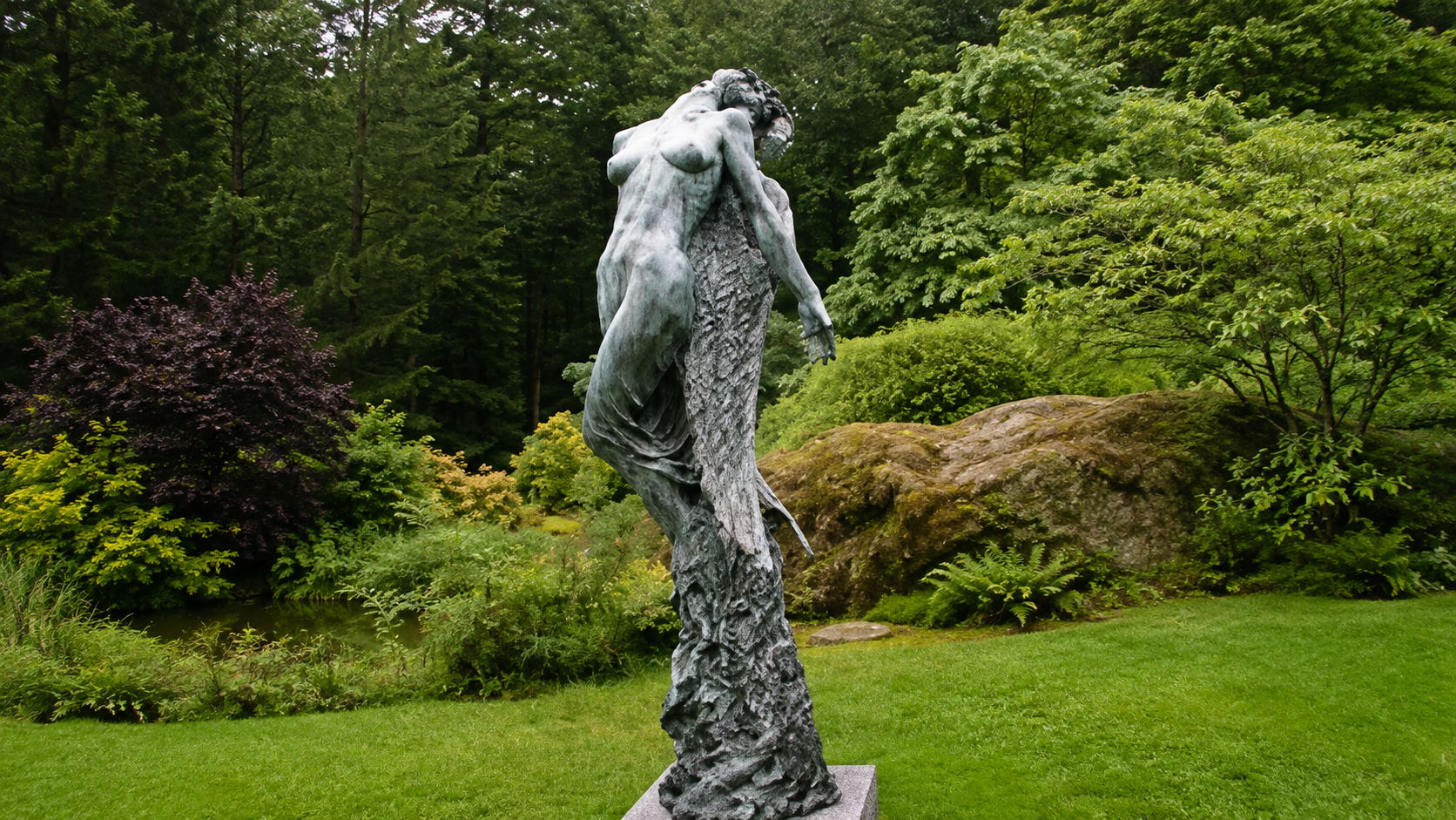 Bronze sculpture of two intertwined women in a lush, green garden with trees and shrubs.