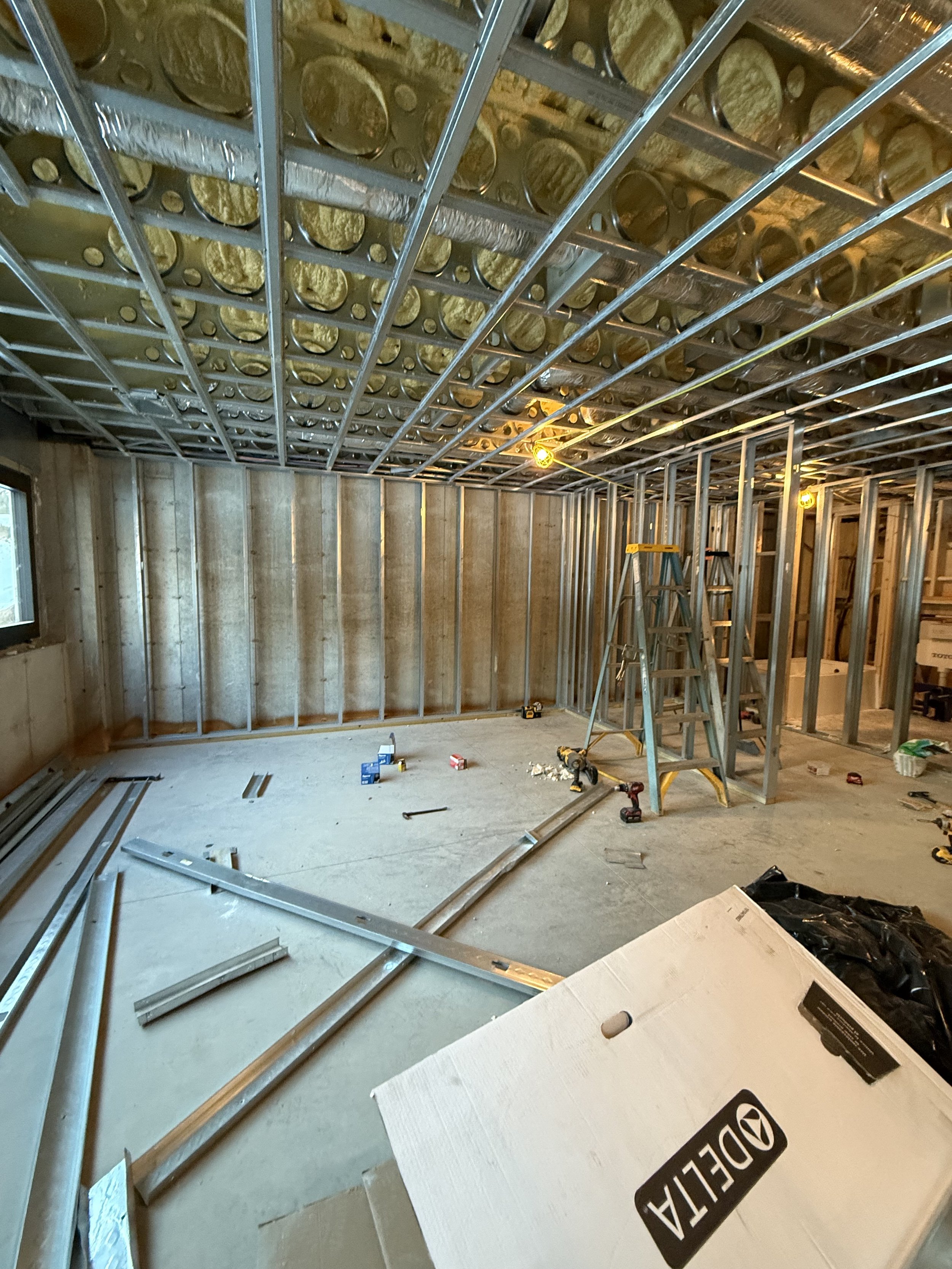 Interior room under construction with metal framing for walls and ceiling, ladders, tools, and packaging materials scattered around.