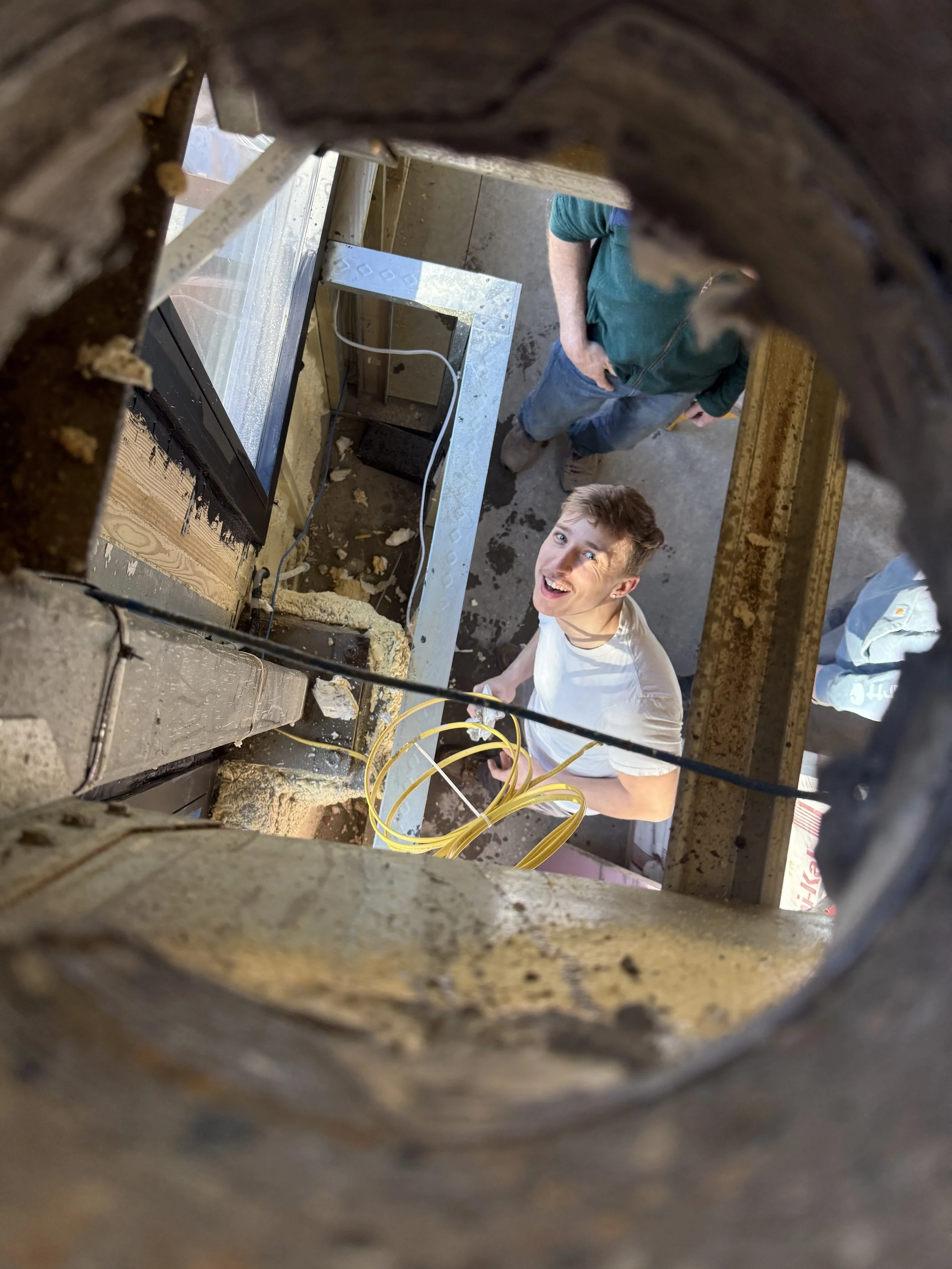 View from inside a hole or pipe looking up at two people standing in a construction or renovation site. One person is wearing a green shirt and the other is wearing a white shirt. The scene includes construction materials, wires, and framing.