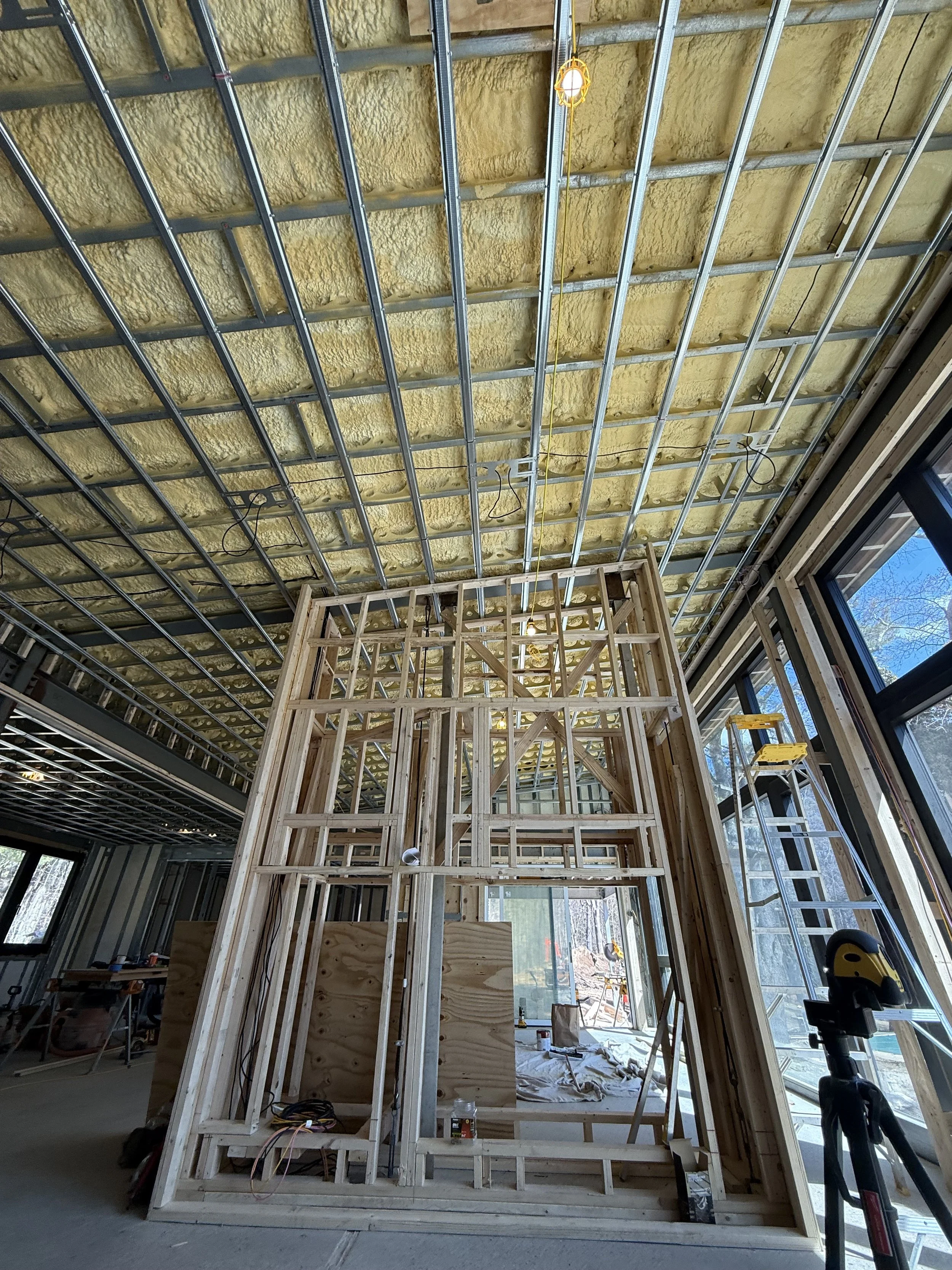 Interior of a house under construction with wooden framing and ceiling insulation, large windows, and construction tools visible.