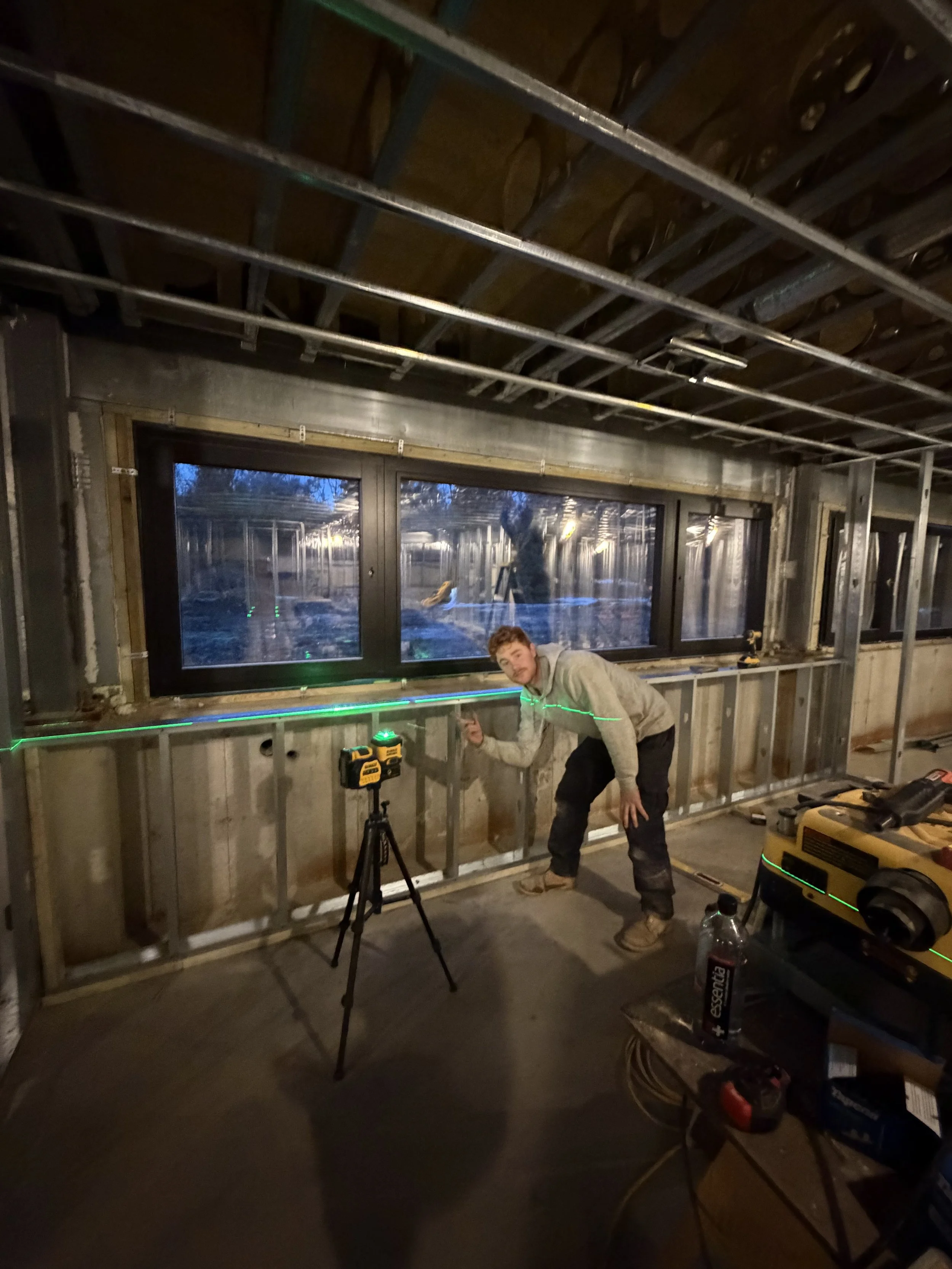 A person working on a construction project inside a building at night, with exposed metal framing, a window, and tools including a drill, a laser level, and a water bottle on a work table.