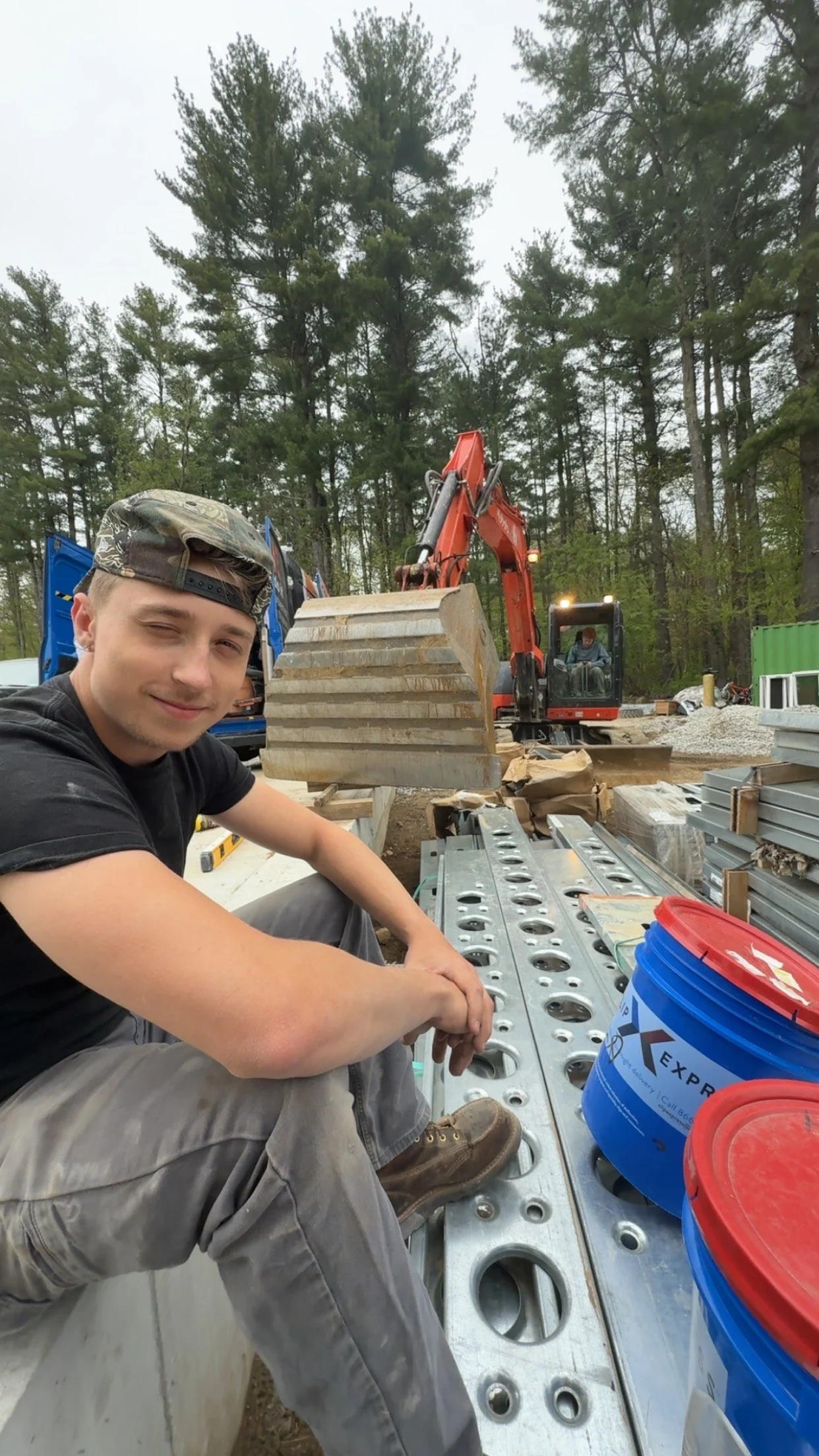 A young man sits on a construction platform at a worksite, smiling and looking at the camera. In the background, an orange excavator and another worker are visible, surrounded by building materials and a wooded area.