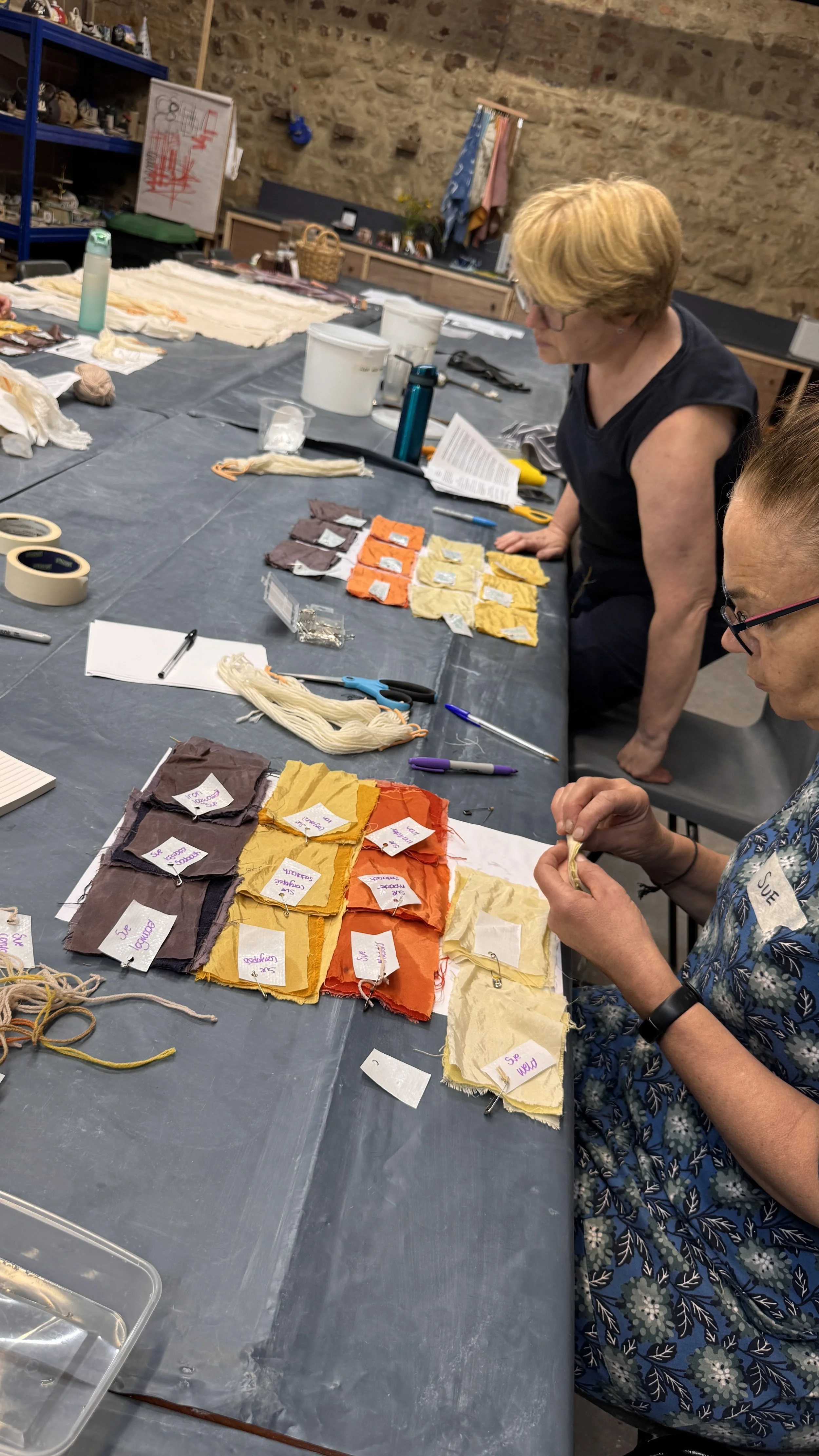 Two women working at a table covered with fabric swatches arranged by color, along with scissors, pens, and small labeled papers. The room has a brick wall background, shelves with various items, and a whiteboard.