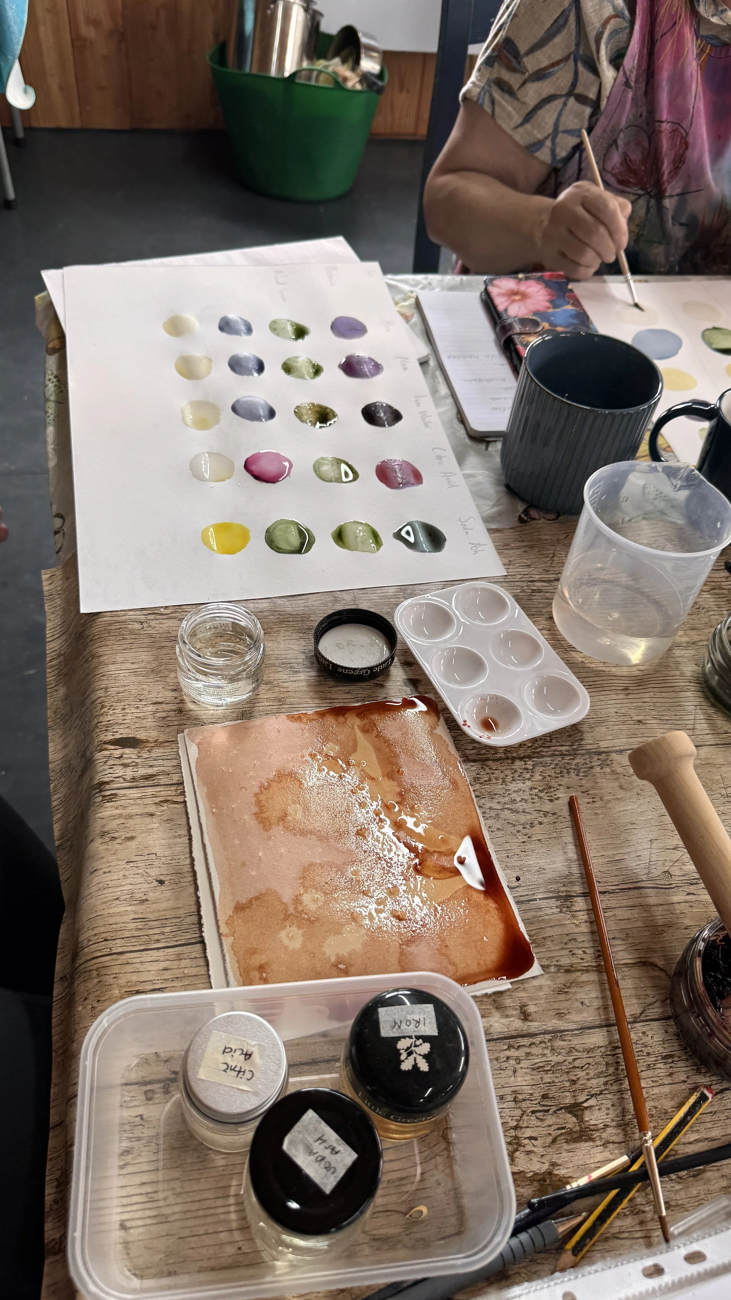A person painting watercolor on paper at a wooden table, with various paint colors, brushes, and containers nearby. The person is partially visible, wearing a floral shirt or apron.