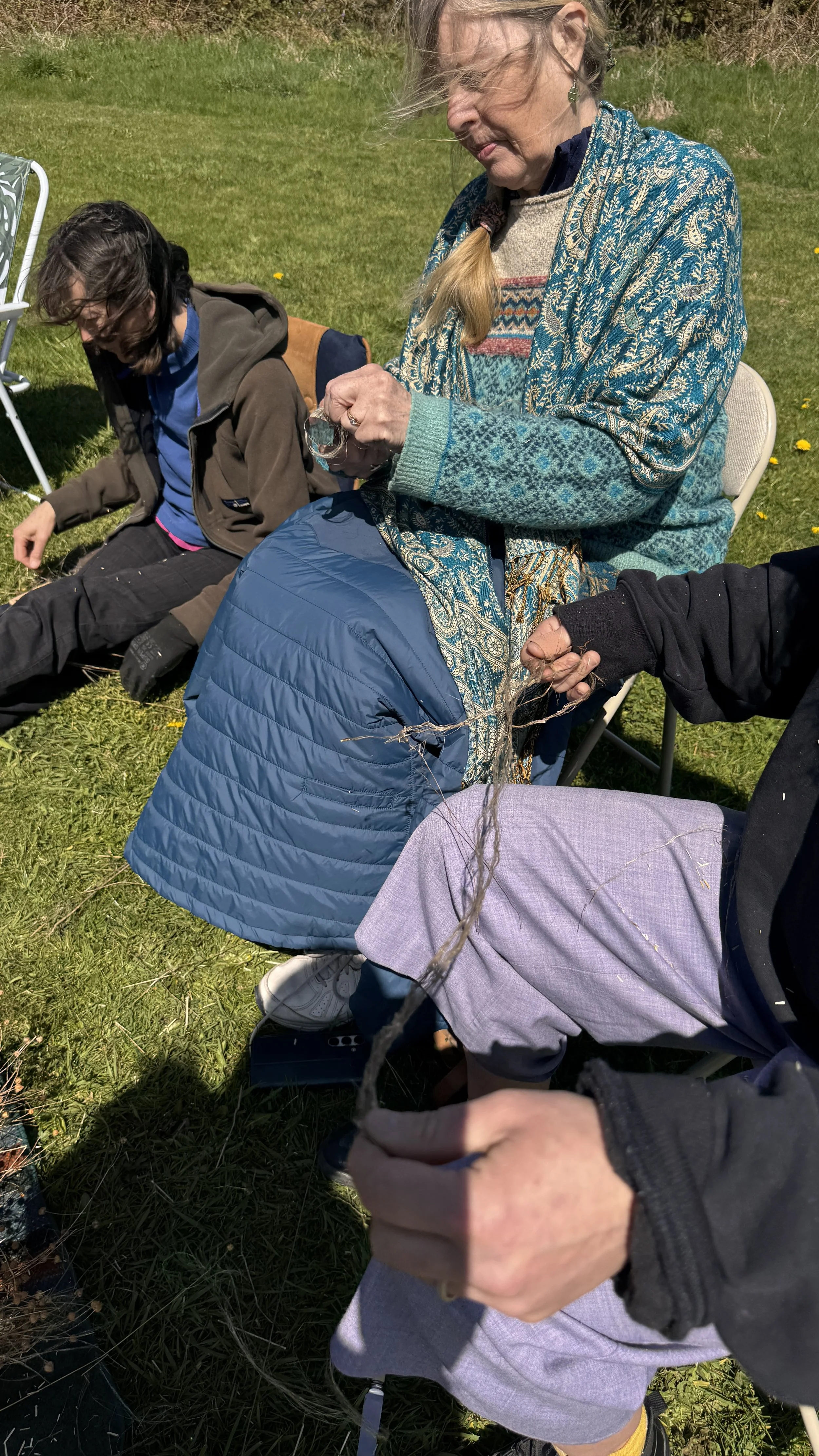 Two people sitting on grass outdoors, one woman wearing a colorful sweater and a teal-patterned shawl, the other person with long hair and a brown jacket, holding a piece of string or plant. The background shows green grass and some small yellow flow