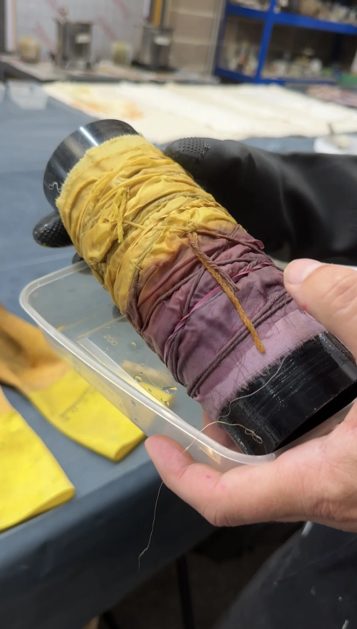 A person holding a black cylindrical object wrapped in yellow and purple cloth, with the cloth tied with a yellow string. The background shows a workspace with yellow bags and blurred shelves.