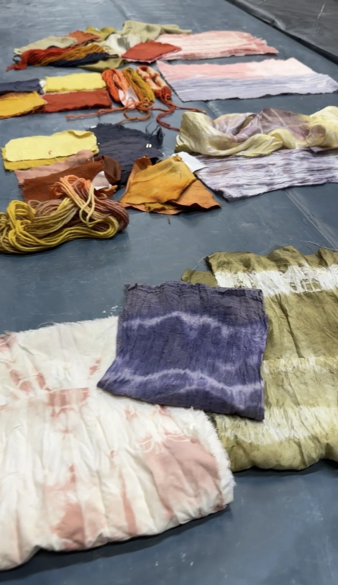 Various pieces of fabric, yarn, and garments are spread on a gray surface, with some yarns tangled together and some pieces folded or crumpled.