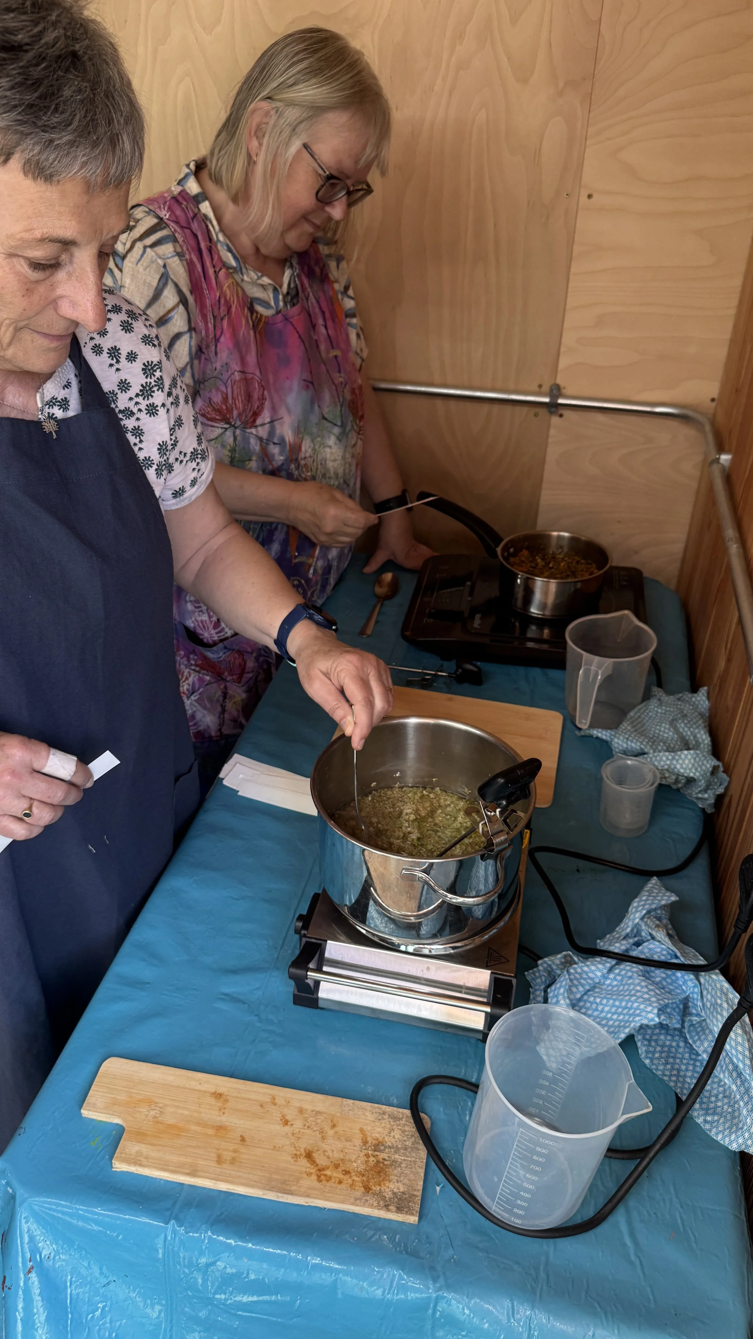 Two women cooking food on a stovetop in a kitchen with wooden walls, with various cooking utensils and containers on a blue tablecloth covering the table.