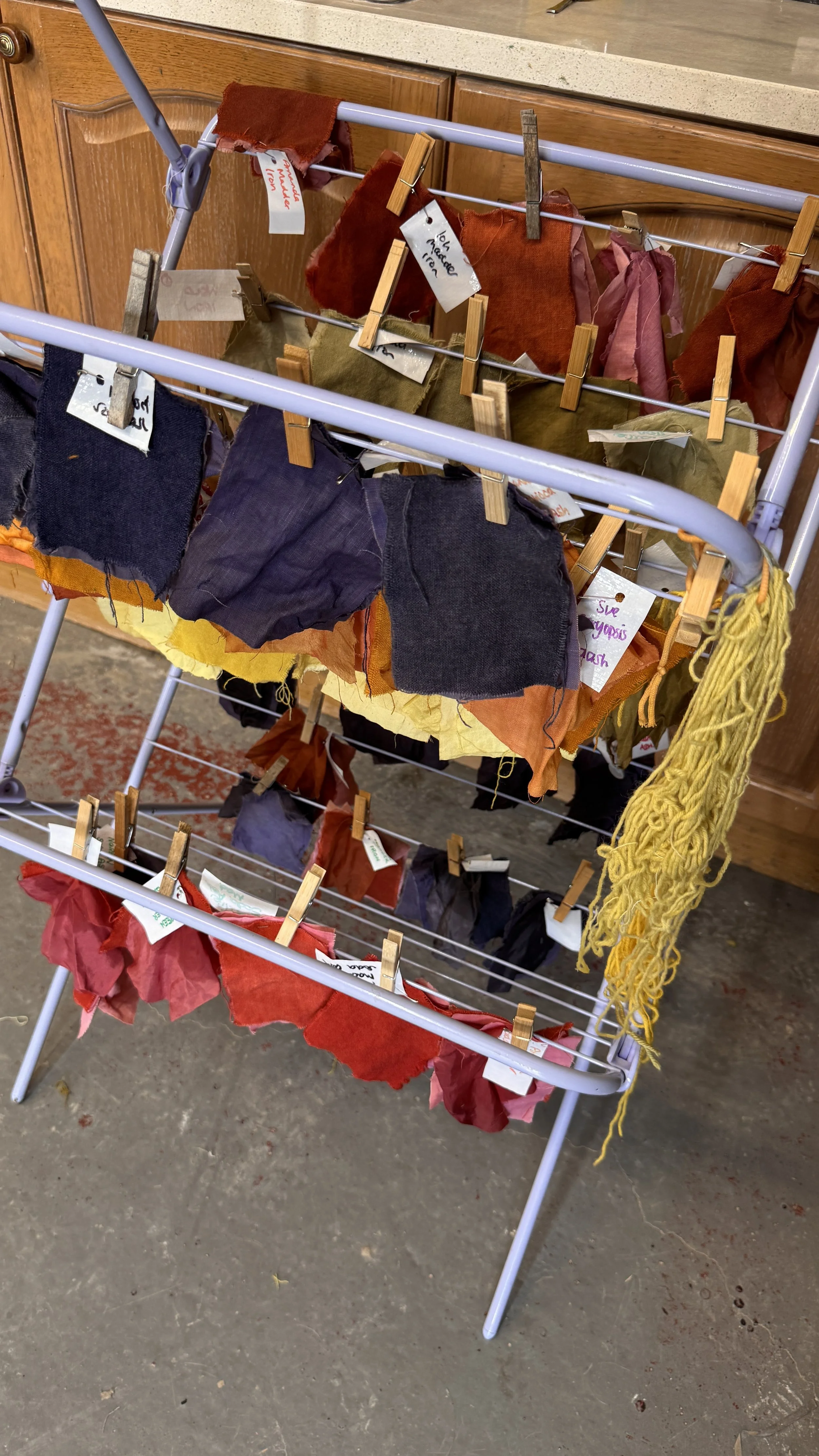 A white clothing rack with three shelves holding fabric samples and swatches in red, orange, yellow, and dark colors, secured with wooden clothespins, in a room with a wooden cabinet and concrete floor.