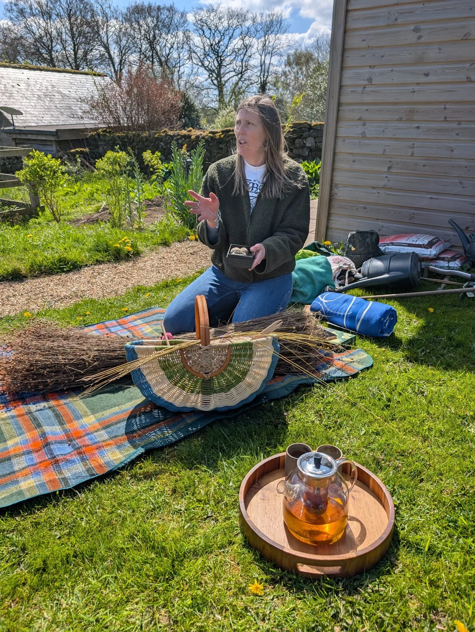 A woman sitting on a colorful blanket in a garden, holding a small box, with various gardening and picnic items around her, including a glass teapot with tea in a wooden tray on grass, on a sunny day.