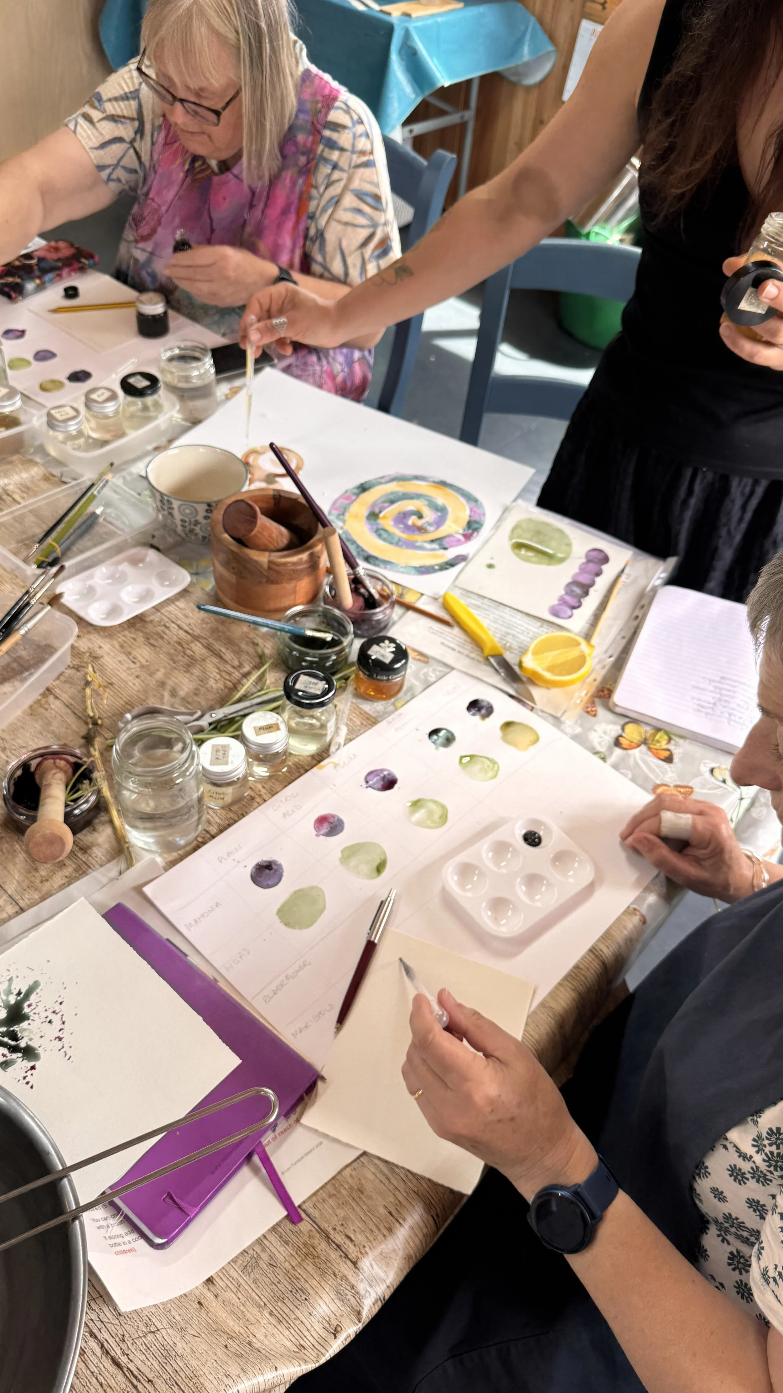 People engaged in watercolor painting and color mixing at a table with art supplies, sketches, and a work-in-progress watercolor piece.