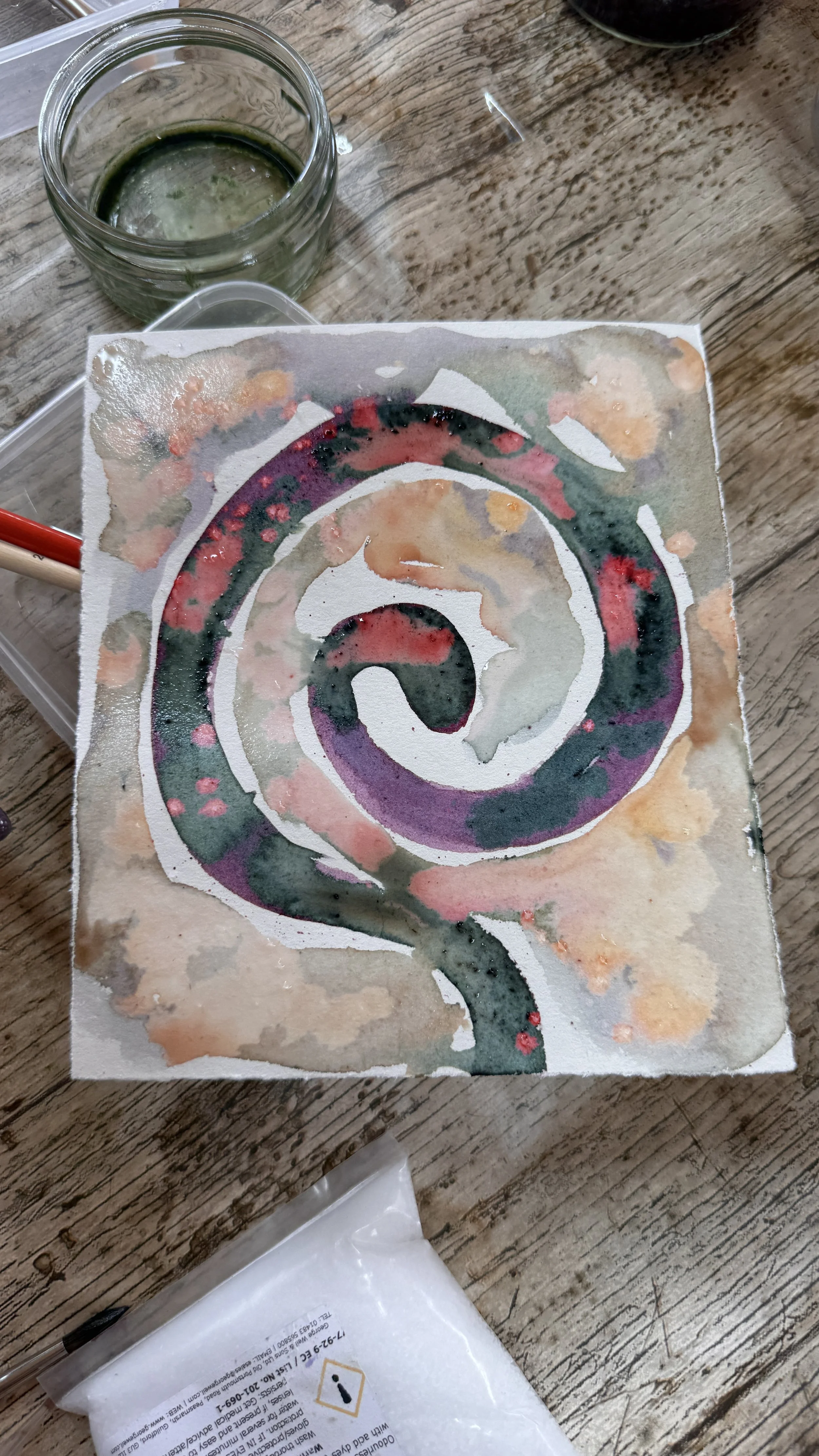 A colorful spiral watercolor painting on paper, with a glass of water and a pen on a wooden surface.