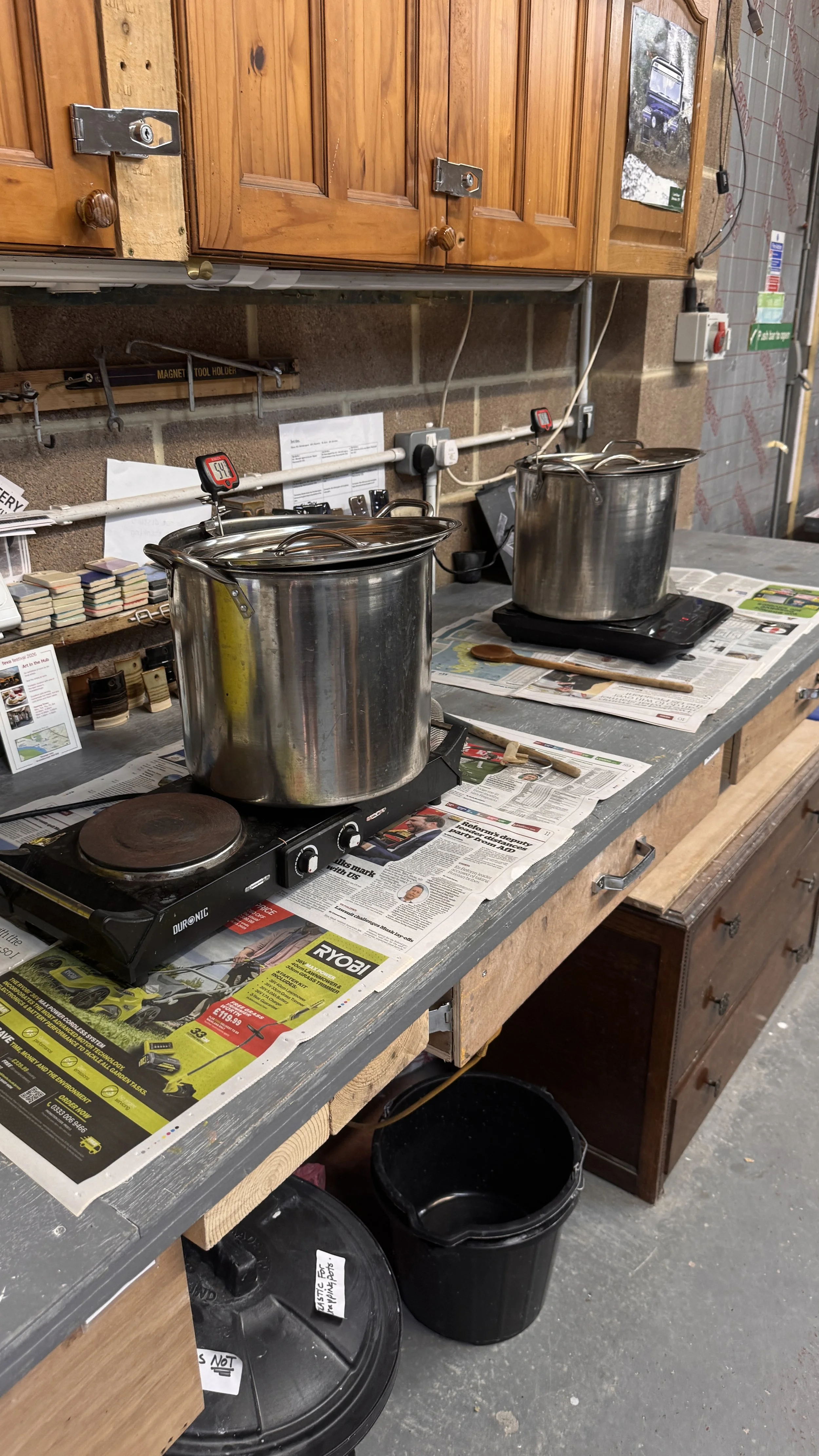Two large stainless steel stock pots on electric stoves on a cluttered wooden workbench covered with newspapers in a workshop or garage with wooden cabinets and various tools and papers.