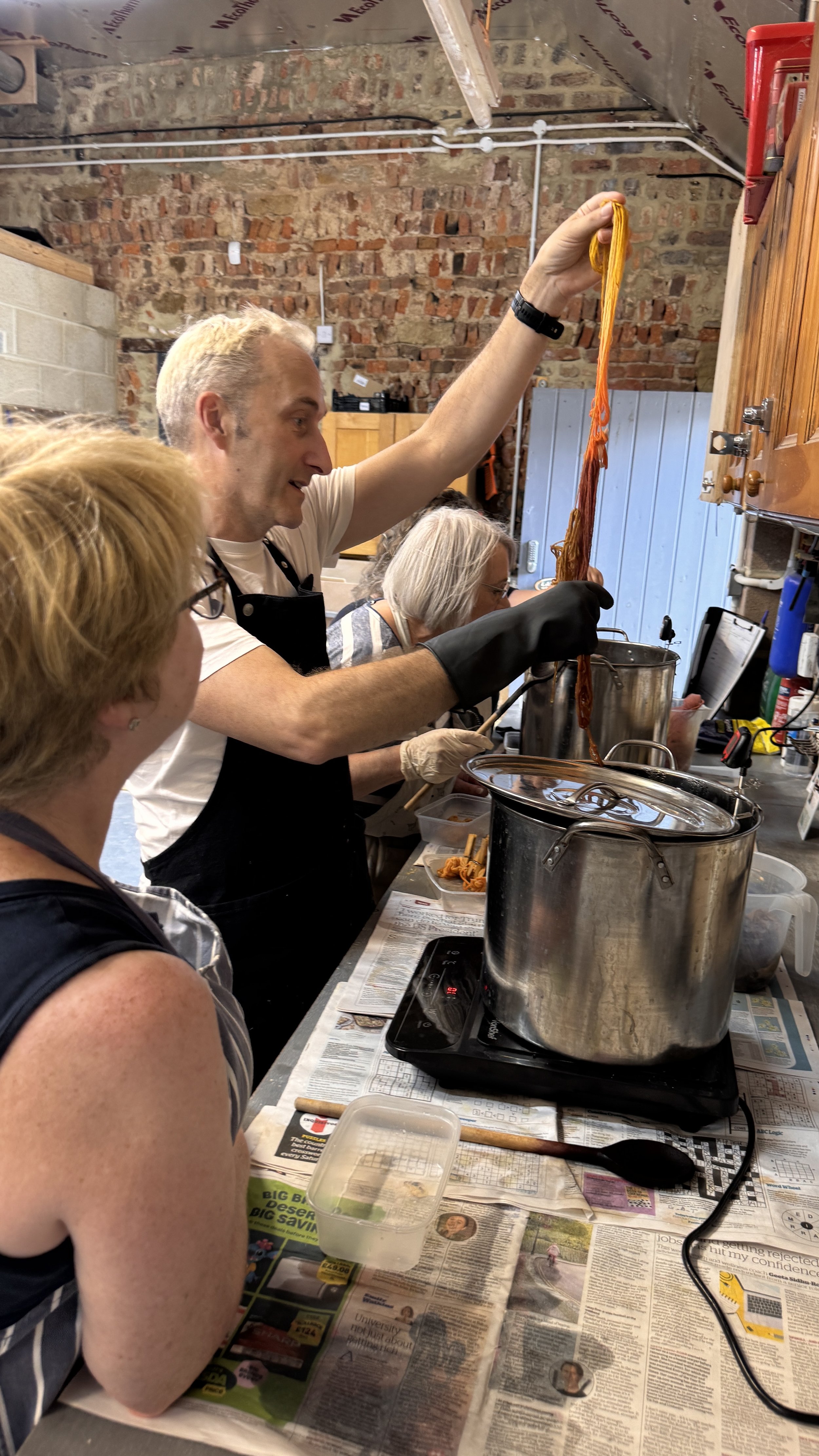 People working in a kitchen, some wearing aprons and gloves, preparing food. The man in the center is holding a bunch of orange, string-like material over boiling pots, possibly yarn or food ingredients. The workspace has a rustic brick wall and a co