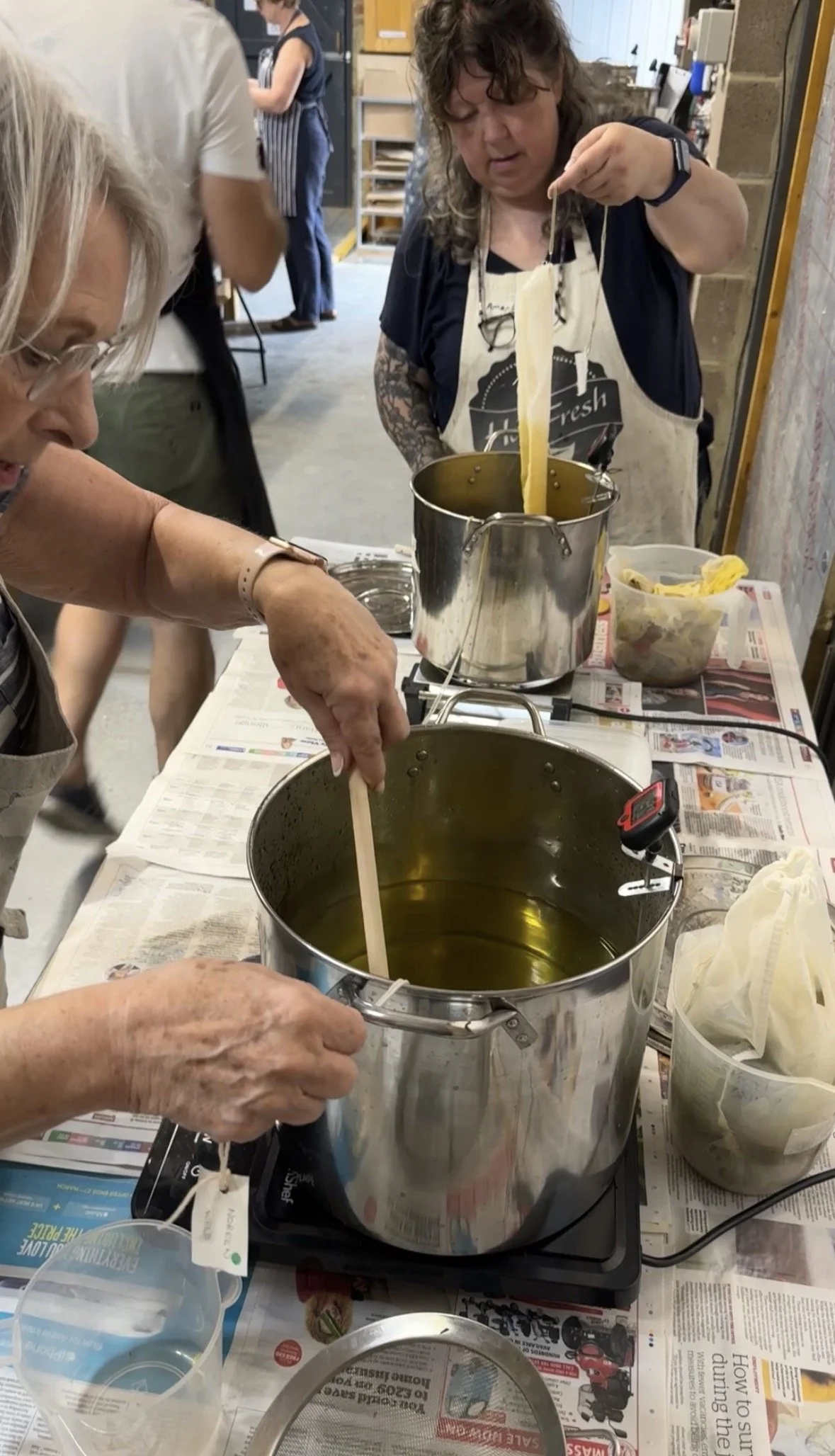 Women making homemade pasta, with one woman pulling a long strip of pasta from a pot and another stirring a pot of hot oil or water, on a table covered with newspapers.
