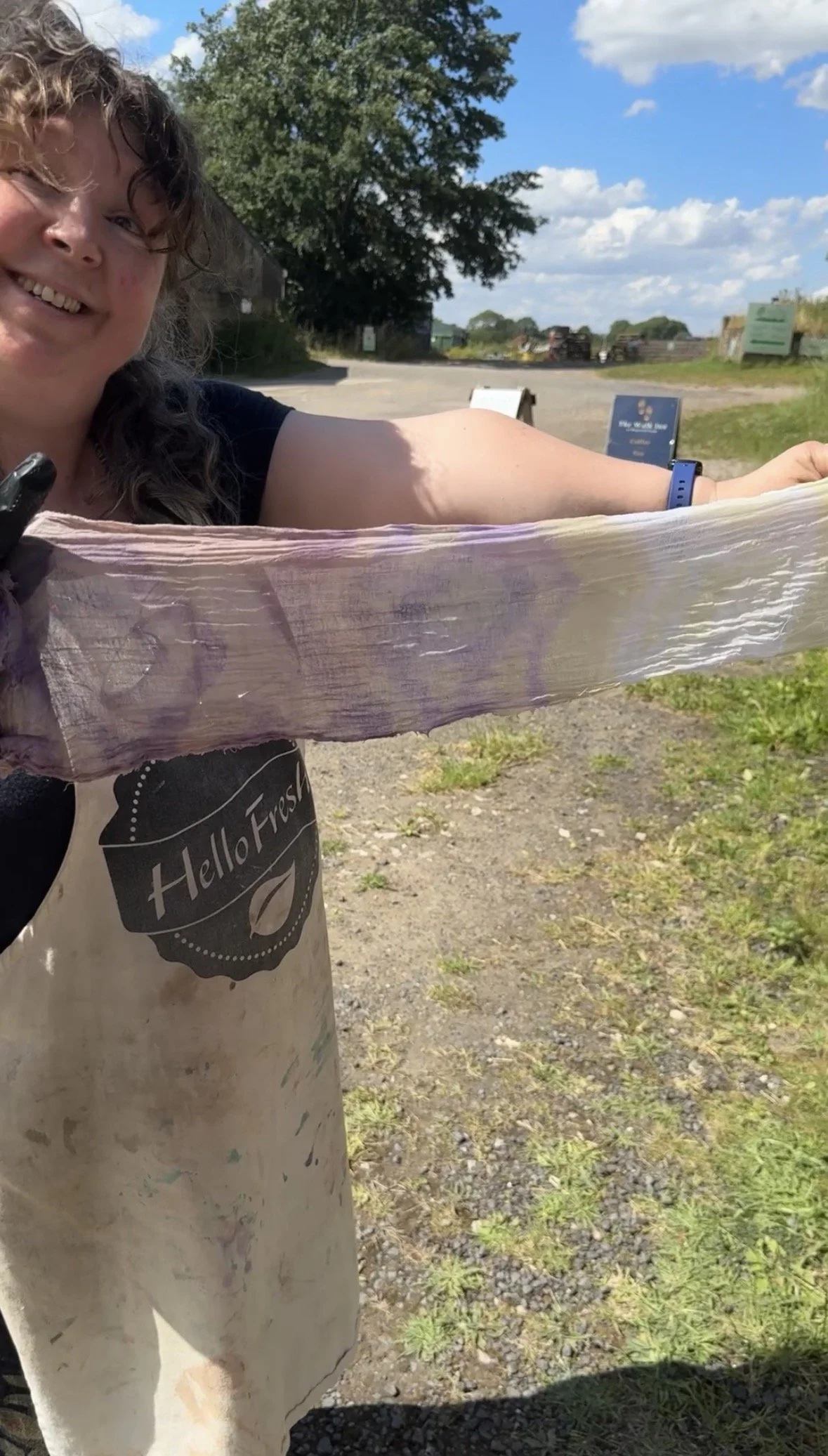 A woman outdoors holding a large, pinkish, fibrous object that appears to be a plant or a vegetable. She is smiling and wearing a black shirt, an apron with a HelloFresh logo, and a sports watch on her wrist. The background features a gravel path, gr