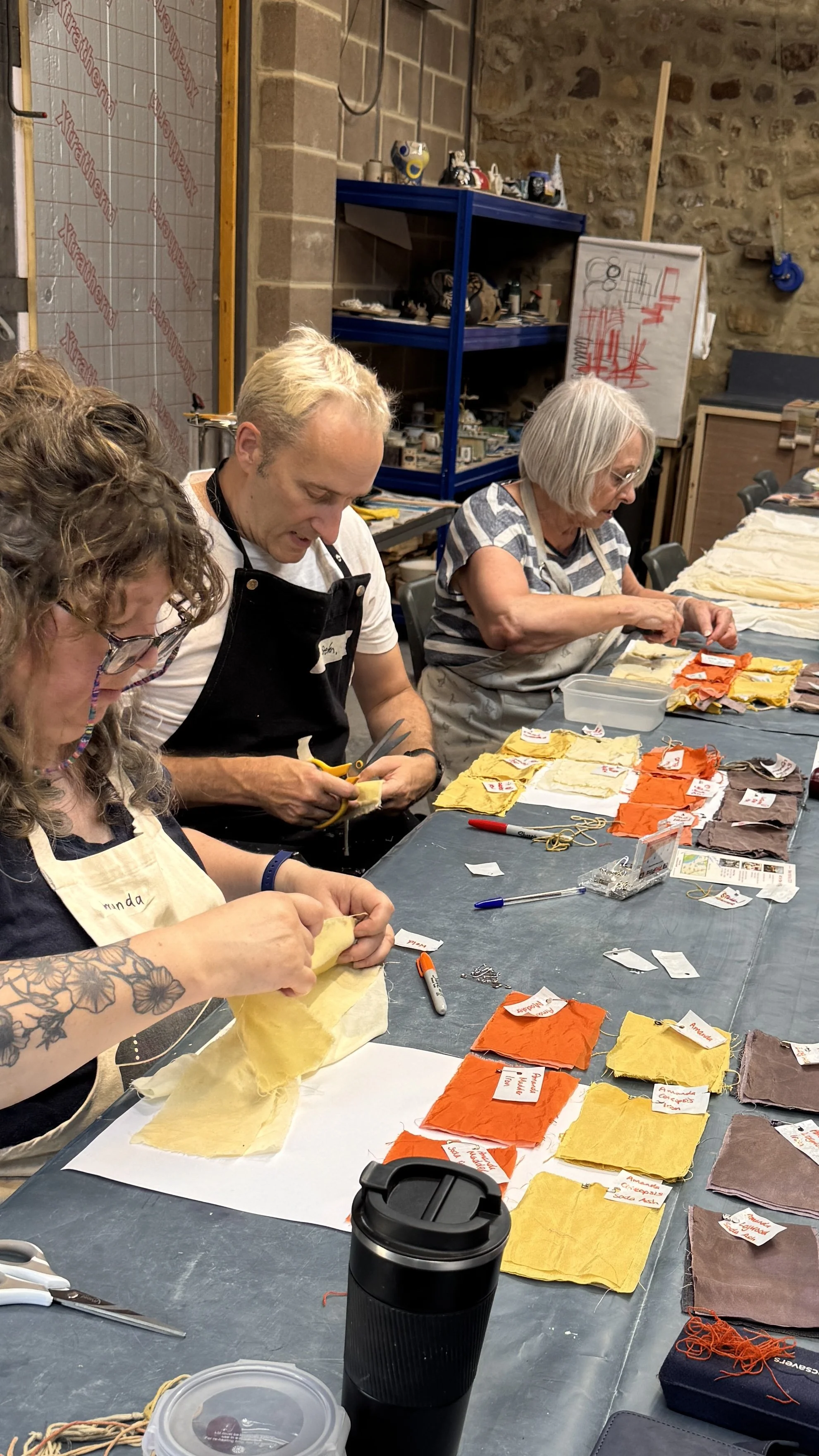 People working with various fabric swatches at a table, with colorful fabric samples labeled with tags, in a workshop setting.