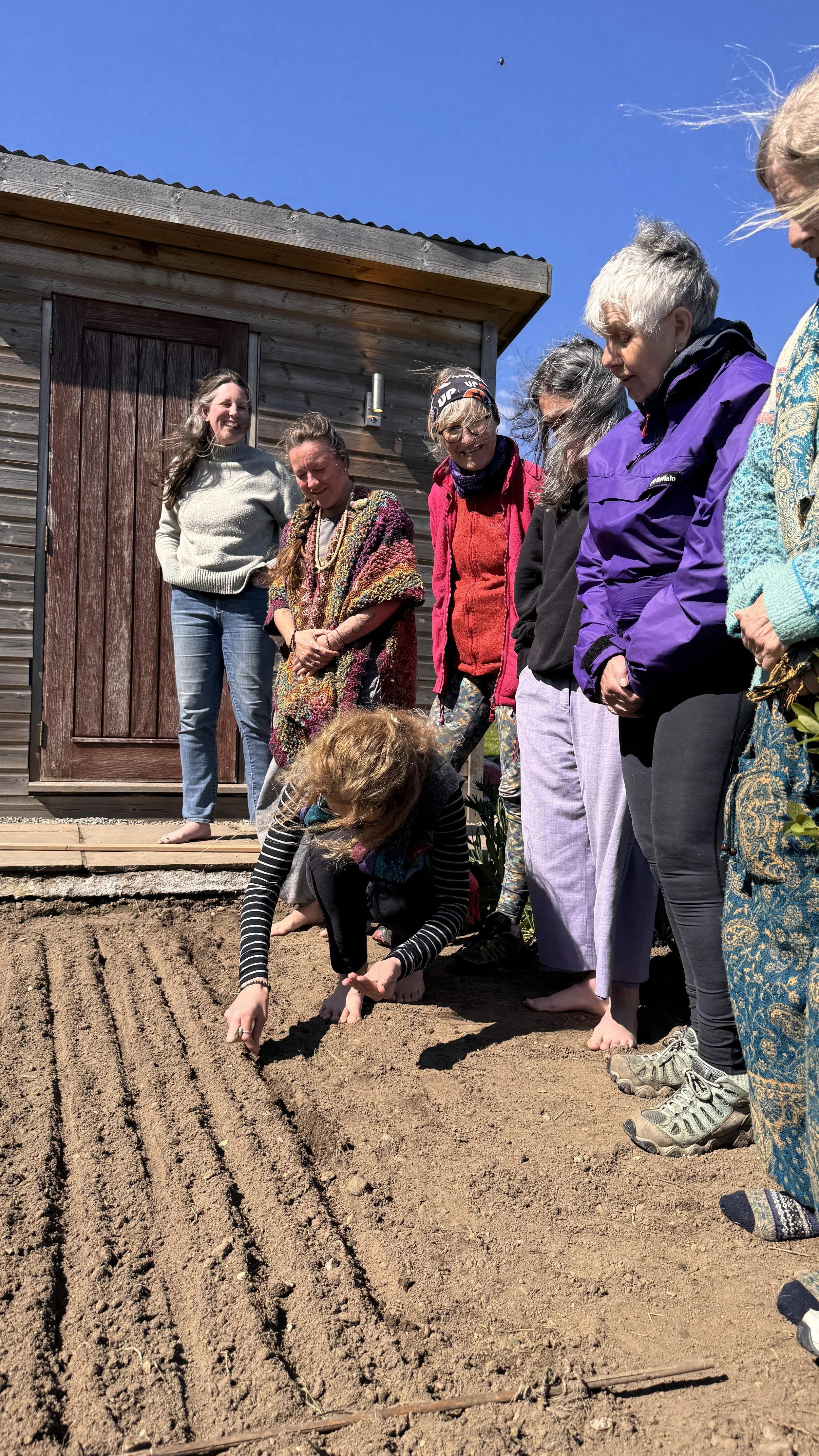 Group of women and children planting or tending to a garden in a yard with a wooden shed, dressed in casual clothing.