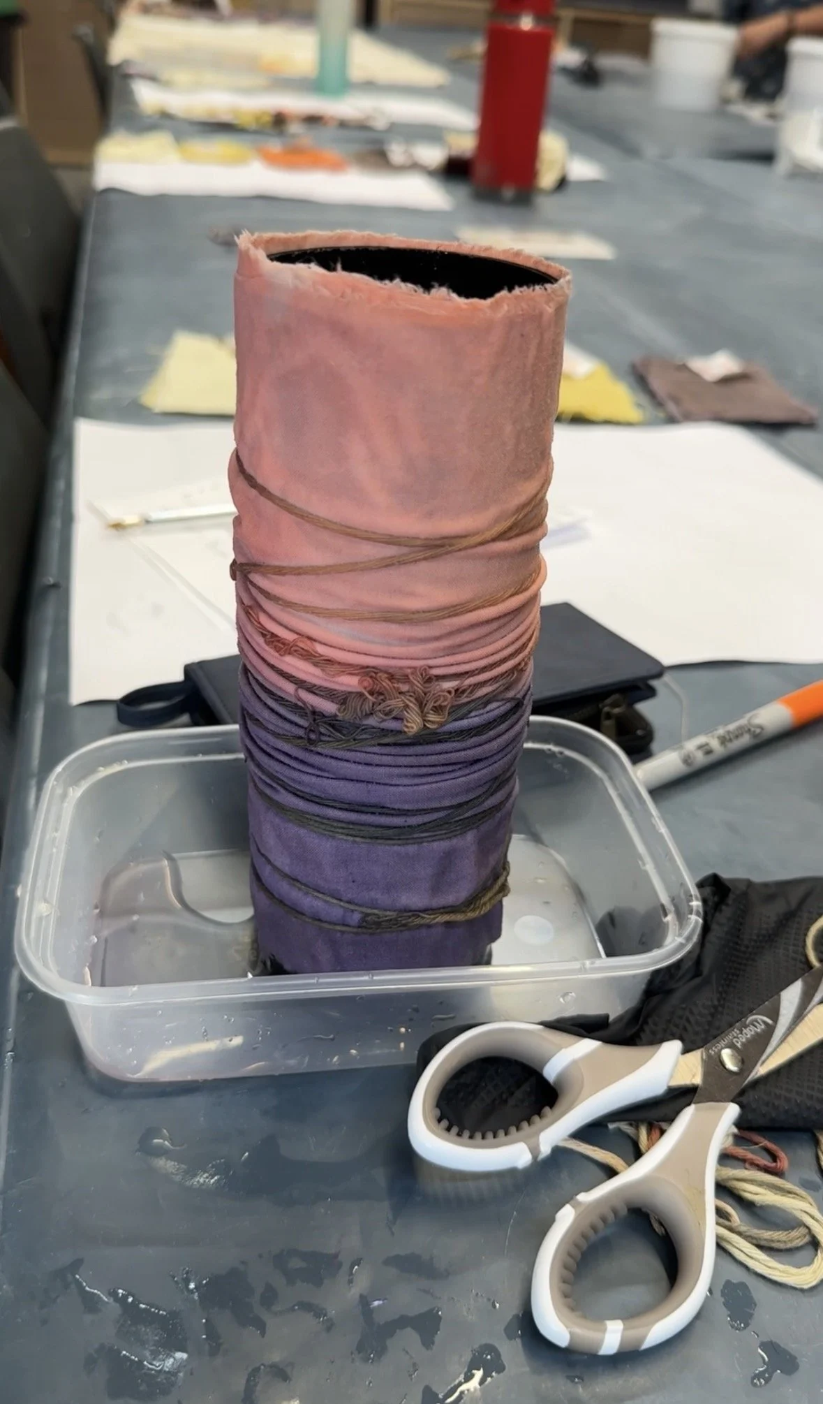 A tall spool of rainbow-colored thread placed in a plastic container filled with water. Scissors and a roll of string are on the table nearby, with a workshop or crafting area visible in the background.