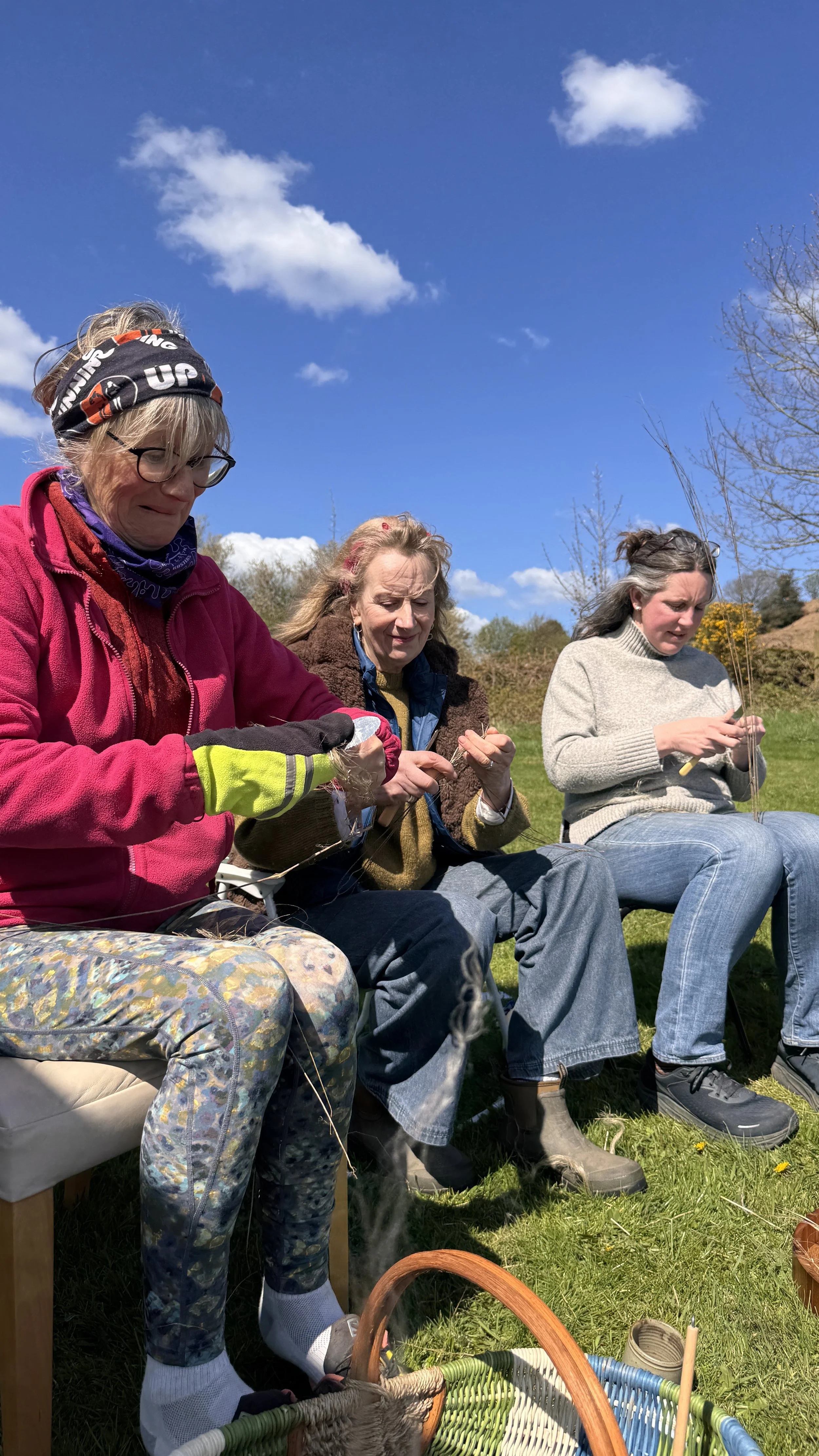 Three women sit outdoors on a grassy field under a blue sky with clouds, practicing basket weaving with natural materials.