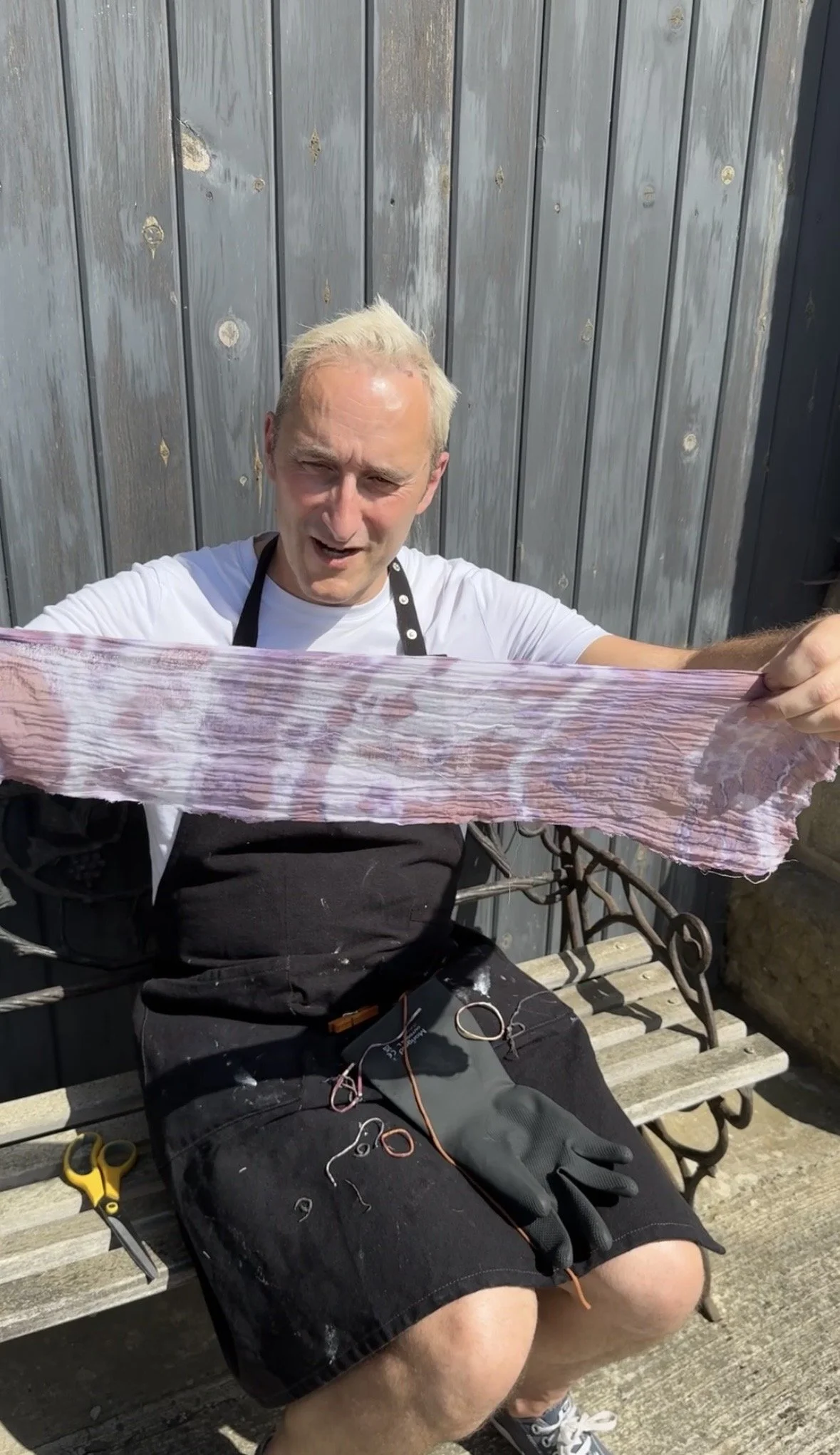 A man sitting on a park bench holding a long strip of raw meat with a wrinkled appearance. The man has short, blond hair, is wearing a white t-shirt, black shorts, and gray sneakers. There are scissors, a pair of glasses, and wires on his lap, with g