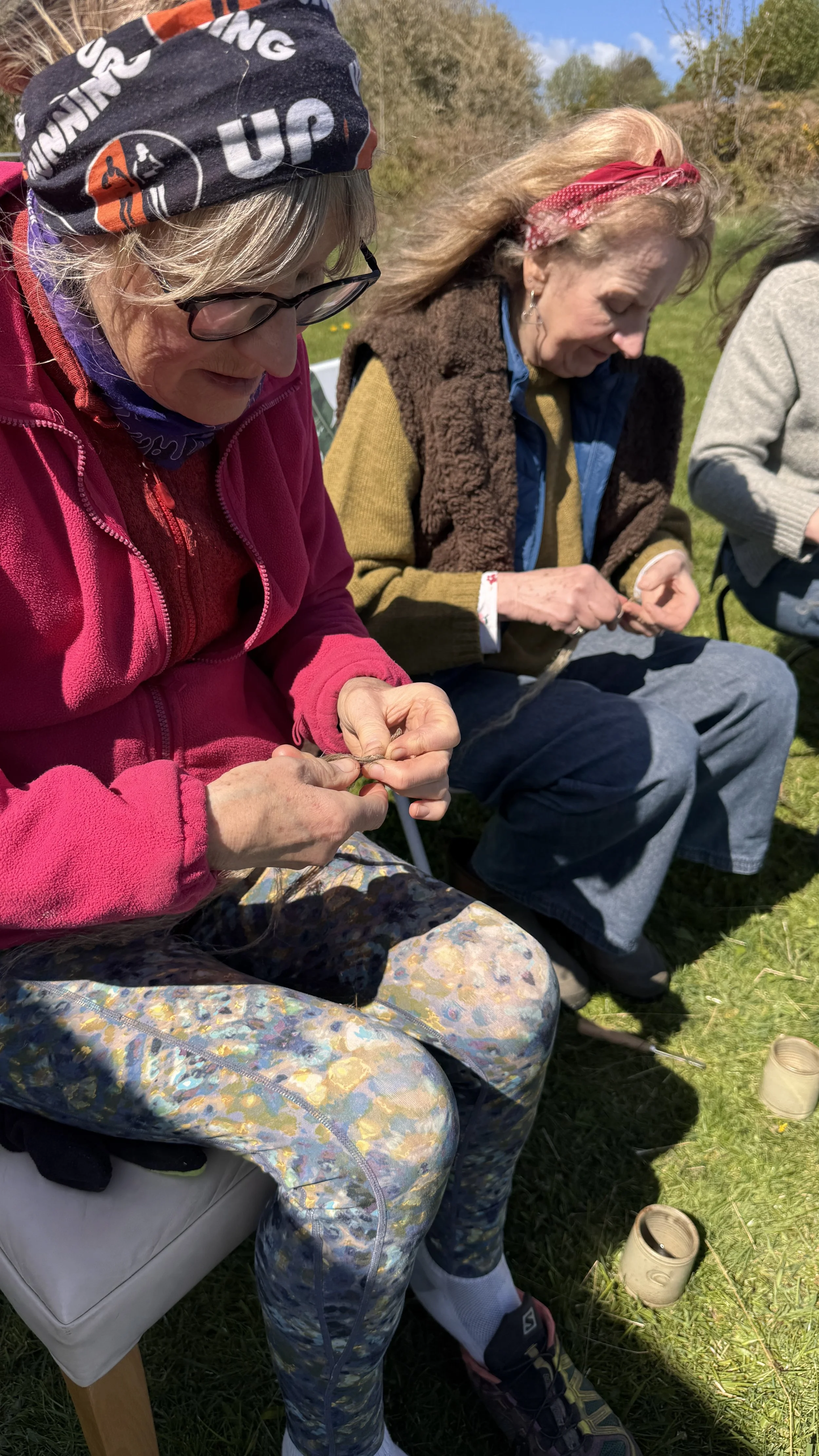 Three women sitting outdoors in a grassy field, engaging in a craft activity with string or yarn. They are dressed in casual, layered clothing suitable for spring or fall, with one woman wearing a black headscarf, glasses, and a bright pink fleece ja