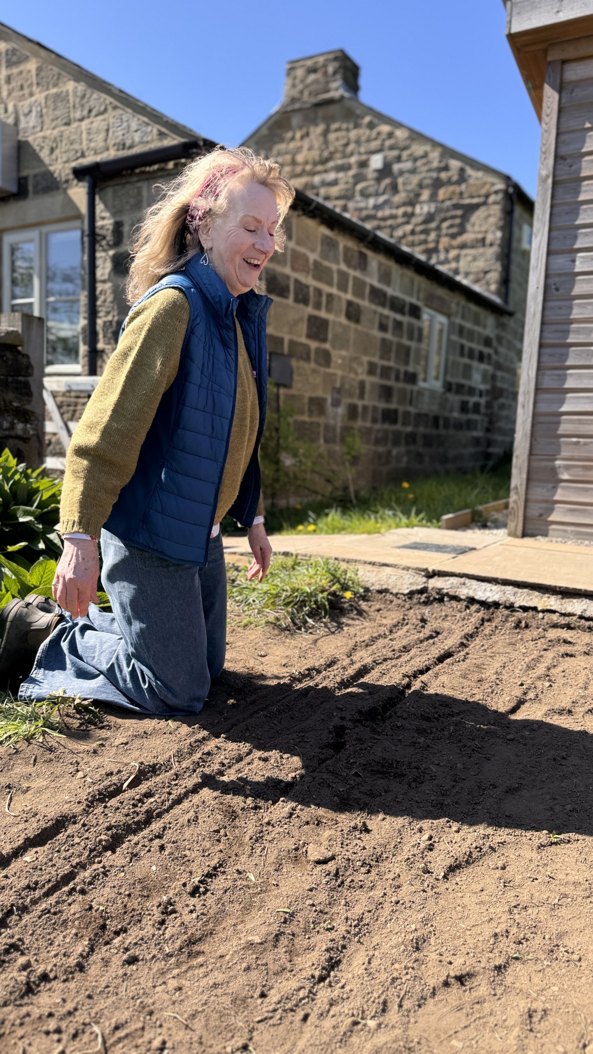 A woman kneeling on her right knee in a garden, smiling, with freshly tilled soil in front of her and a stone house and wooden building in the background.