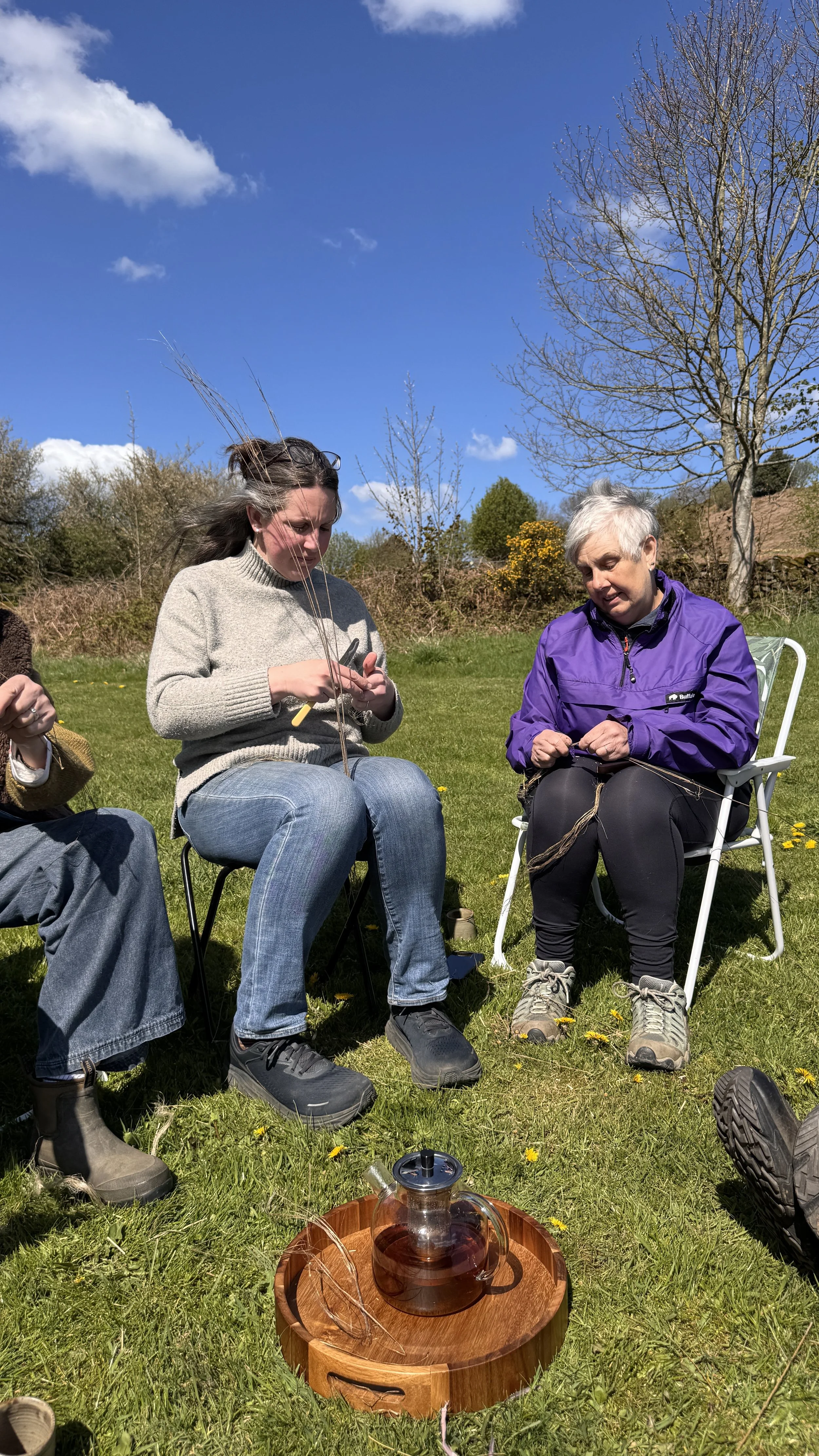 People sitting on chairs outdoors on a grassy field, knitting or weaving with natural materials, with a wooden tray and glass teapot in the foreground.