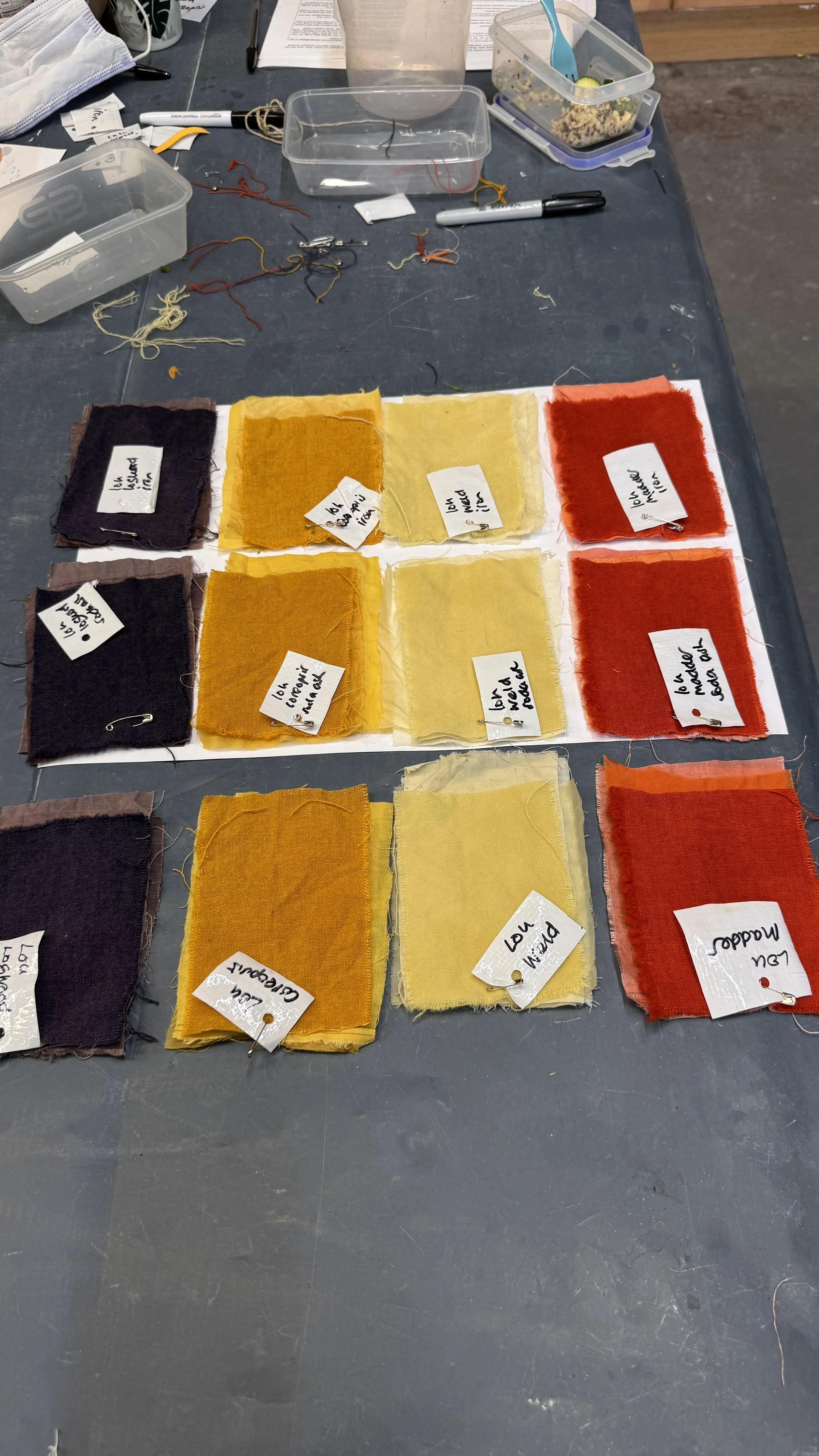 Swatches of fabric in various colors with small handwritten notes attached, arranged on a white sheet on a dark work surface, with sewing supplies and other materials in the background.