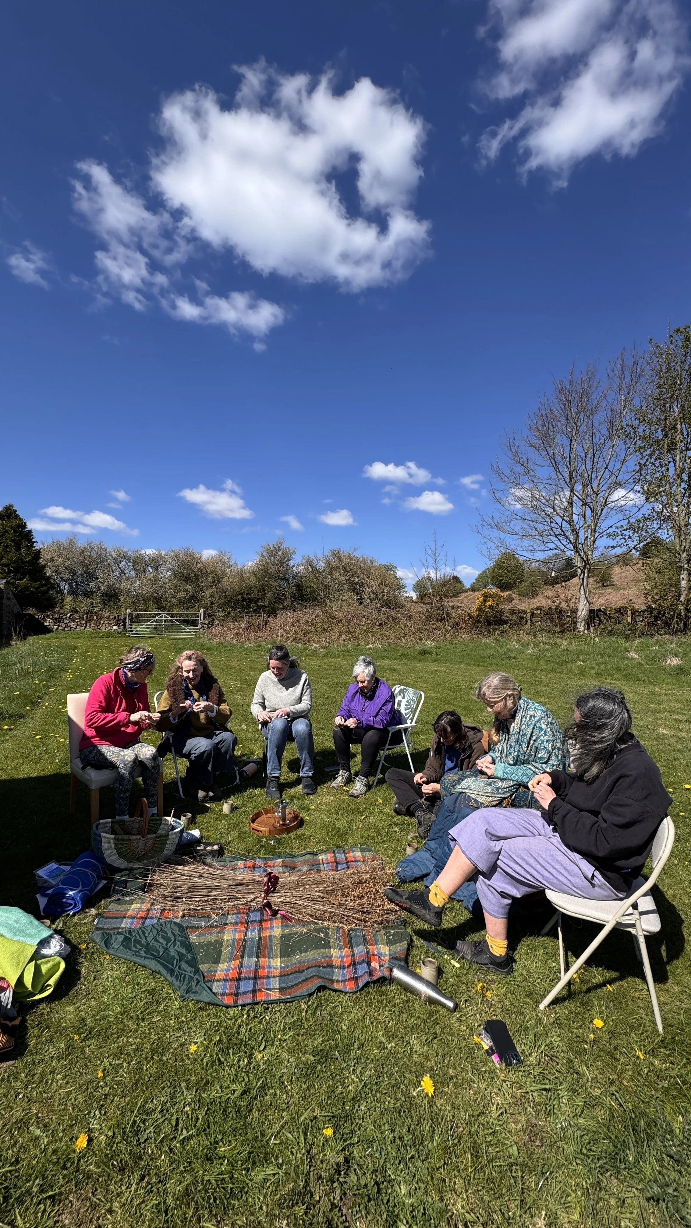 A group of people sitting outdoors on chairs and blankets in a grassy field under a blue sky with clouds. They appear to be engaged in a group activity or discussion, with various items spread out on the ground.