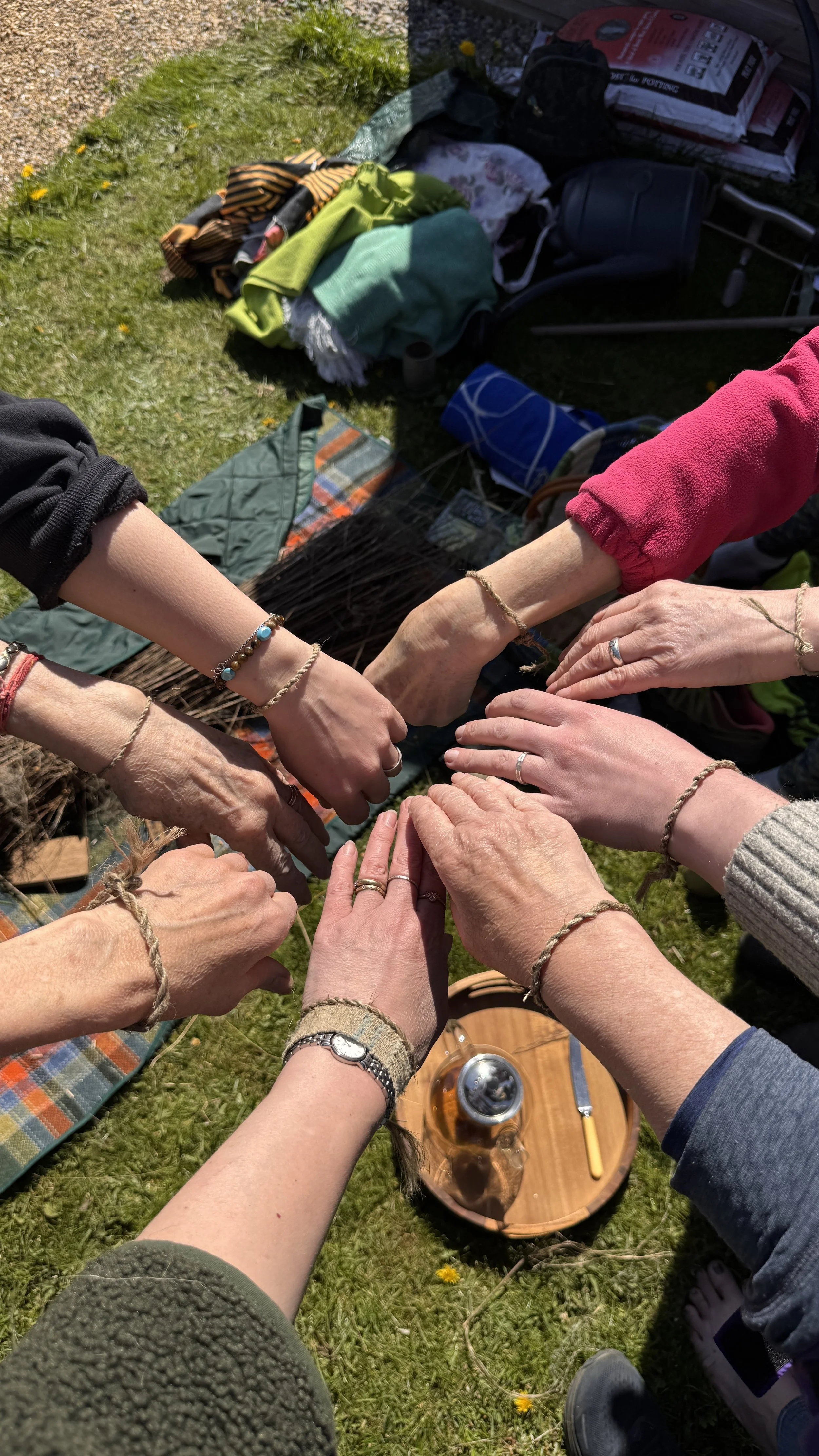 Multiple hands with bracelets and rings joined together in a circle outdoors on grass, with camping supplies and a beverage nearby.