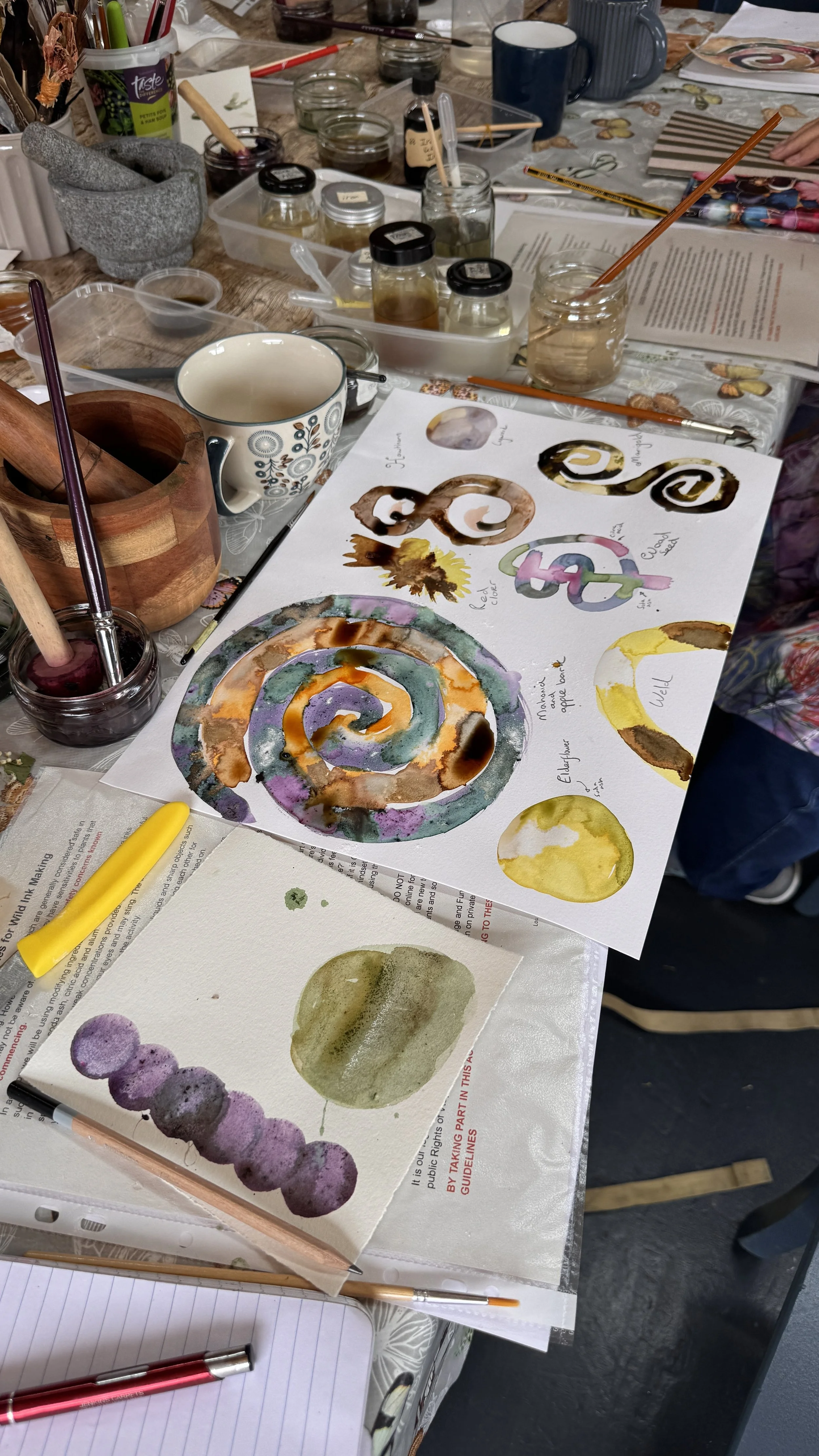 Watercolor artworks of abstract spiral and circular designs, with various colors including brown, purple, yellow, and green, laid out on a cluttered table with jars of water, brushes, and painting supplies.