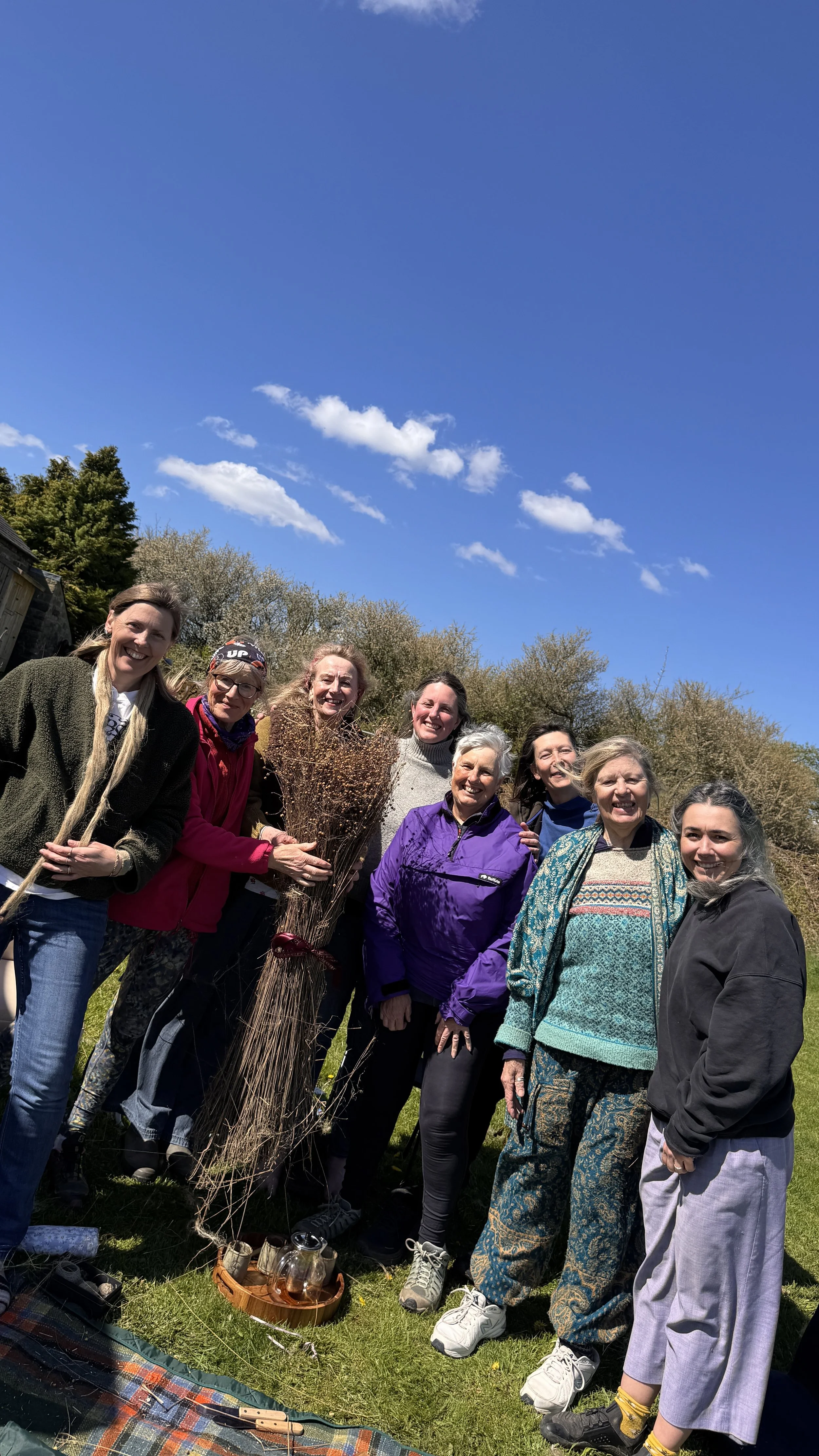 A group of women standing outdoors on a sunny day, smiling, with trees and a blue sky in the background. One woman is holding a bundle of sticks or branches.