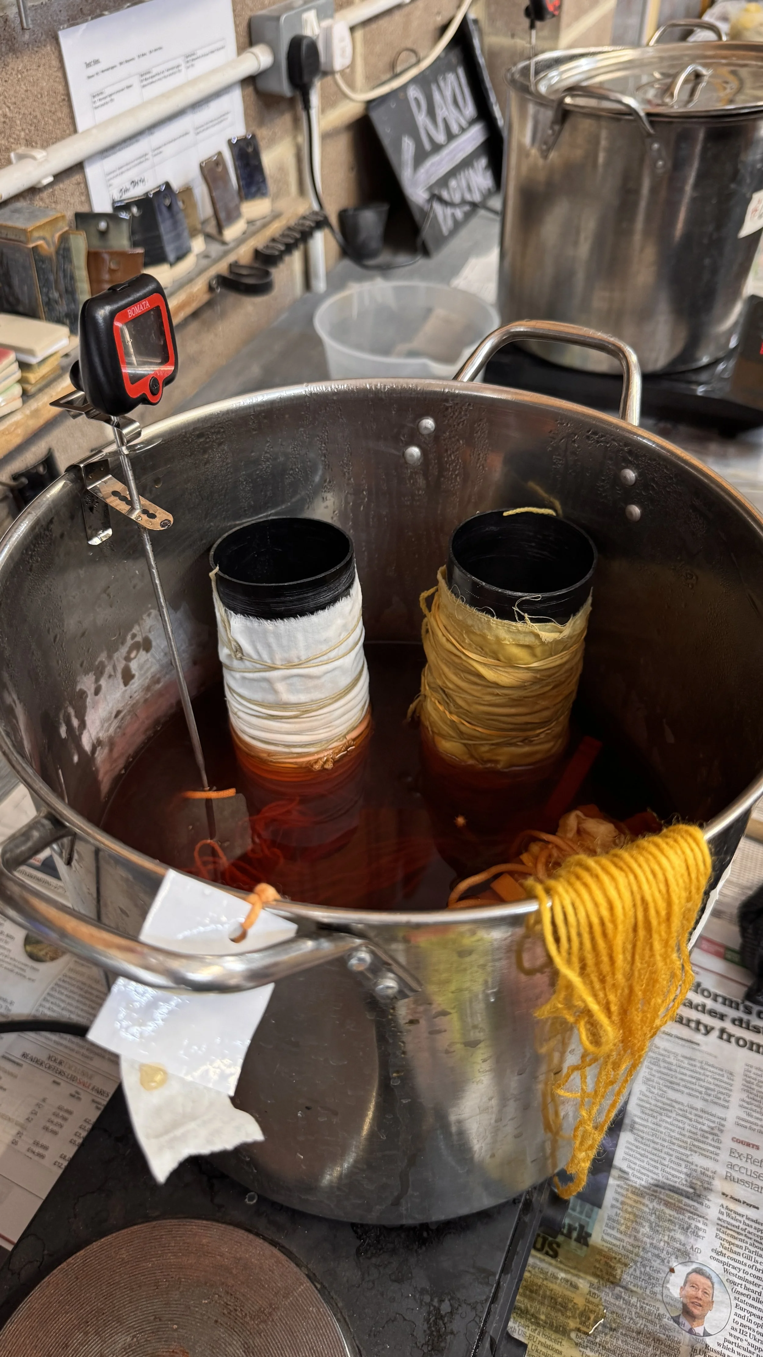 A large stainless steel pot with multiple dyeing socks in colored liquids, with a thermometer inserted into the water, placed on a table in a workshop or dyeing studio.