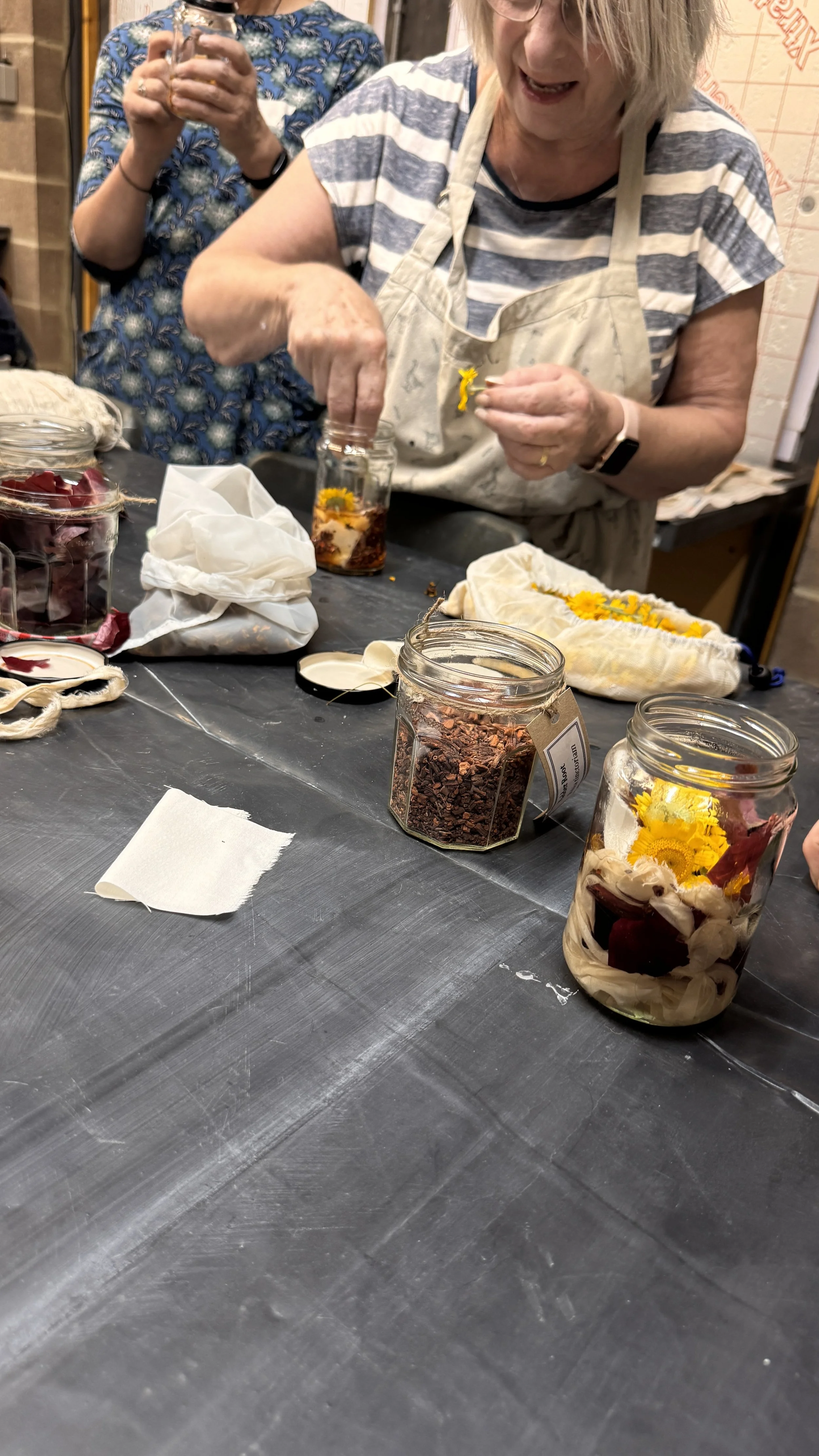 Two women preparing herbal or floral arrangements with jars containing flowers and dried herbs on a table.