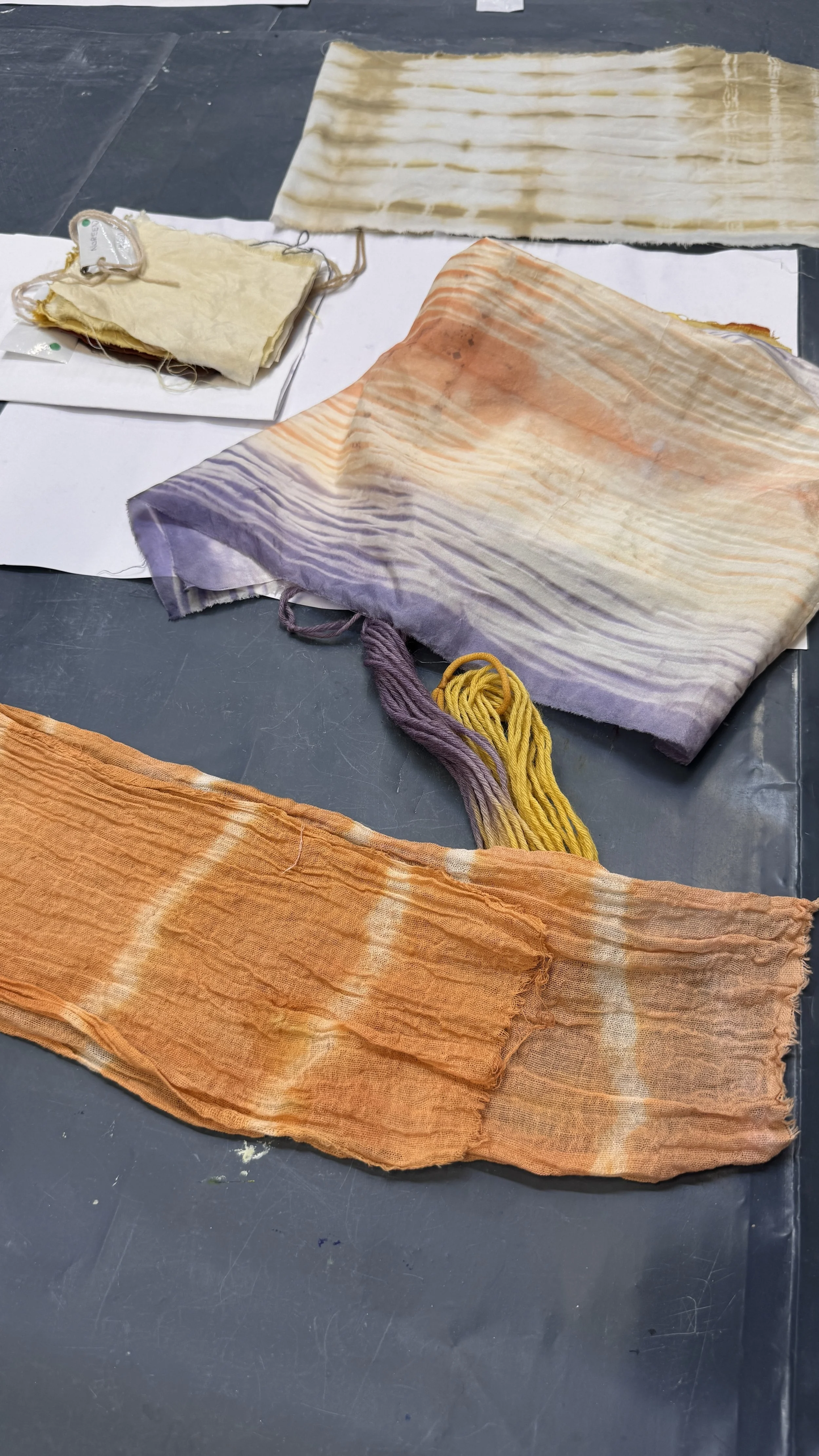 Colorful tie-dye fabric pieces laid out on a dark surface, including a large orange piece, a smaller purple piece with yellow yarn, and a beige piece at the top.