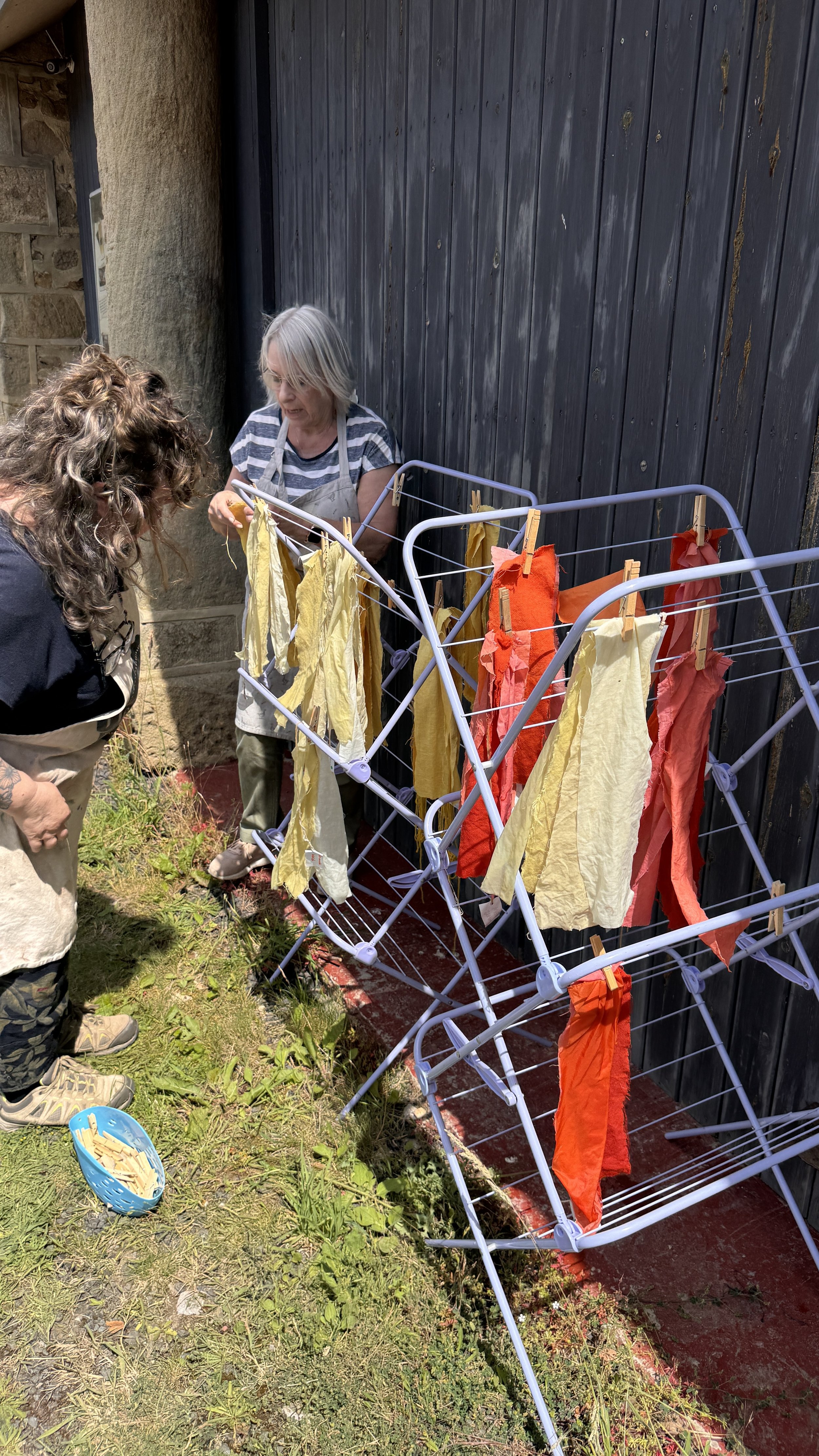 Two women hanging colorful cloths on a clothes drying rack outdoors, next to a stone and wood wall.