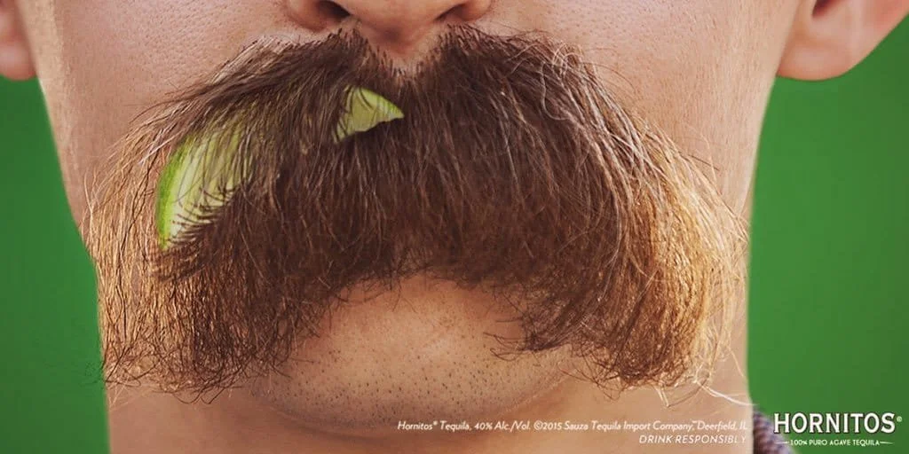 Close-up of a man's face with a large, curly brown mustache, with a lime wedge garnishing the mustache on the left side, against a green background.