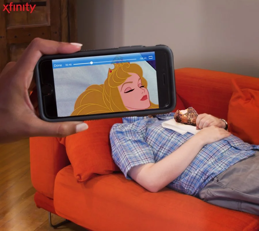 Person lying on a red couch, holding a piece of cake on a napkin on their chest. Another person is taking a photo or video with a smartphone, displaying an animated portrait of a woman with long blonde hair and a crown.