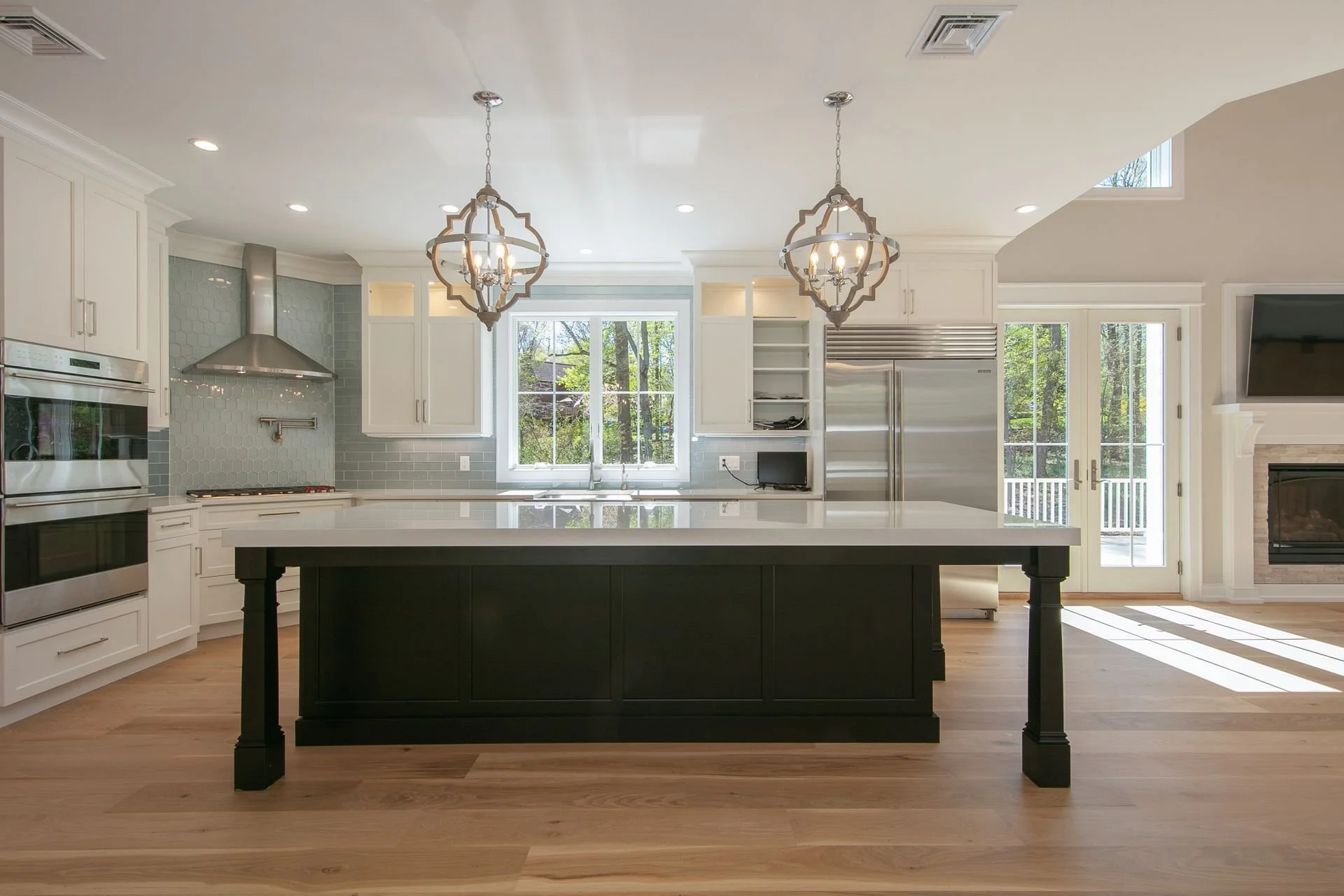 Modern kitchen with white cabinetry, stainless steel appliances, a large kitchen island with a black base, two hanging chandeliers, wooden flooring, large windows, and a glass door leading outside.